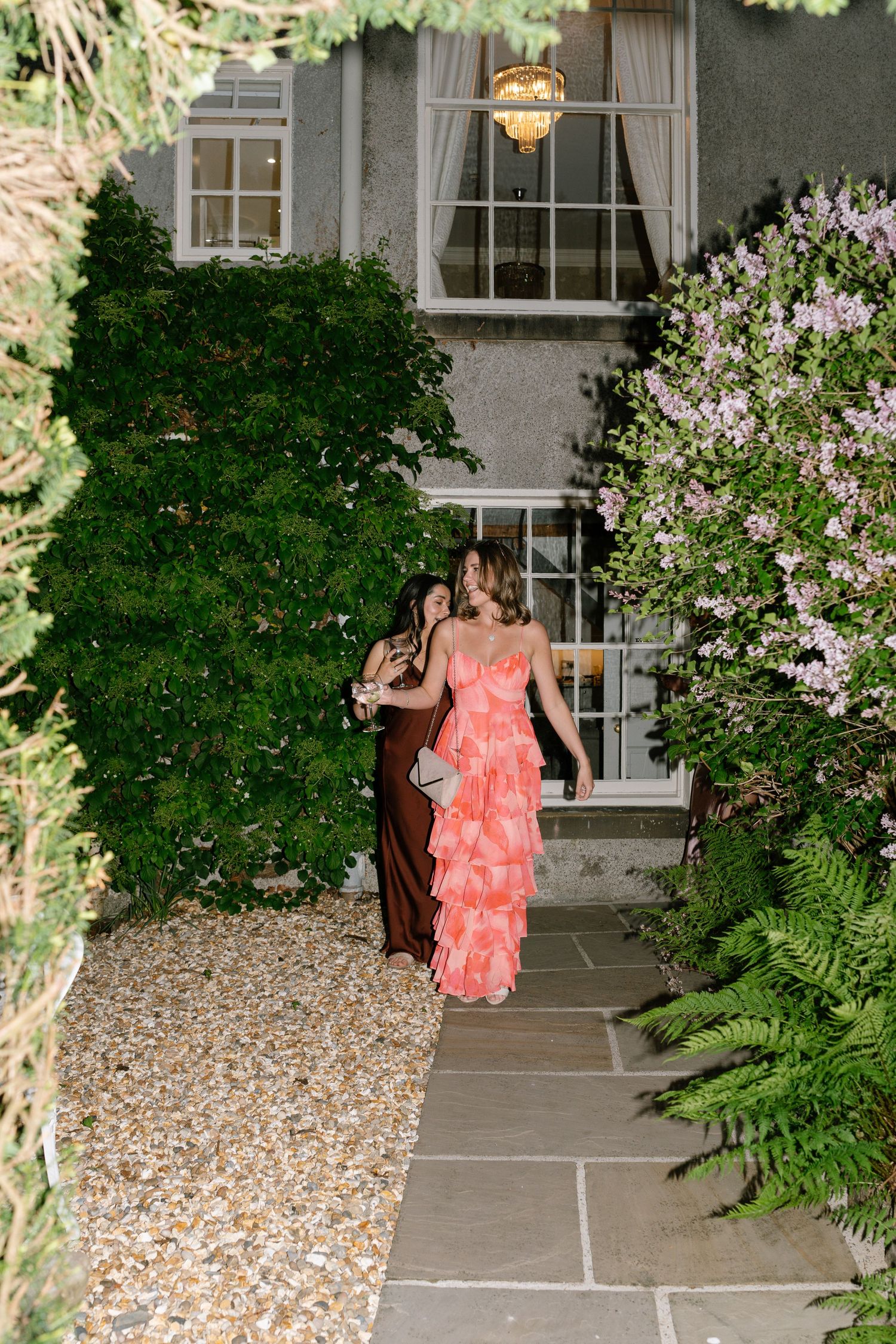 A person in a coral ruffled dress walks along a garden path with blooming flowers and ivy.