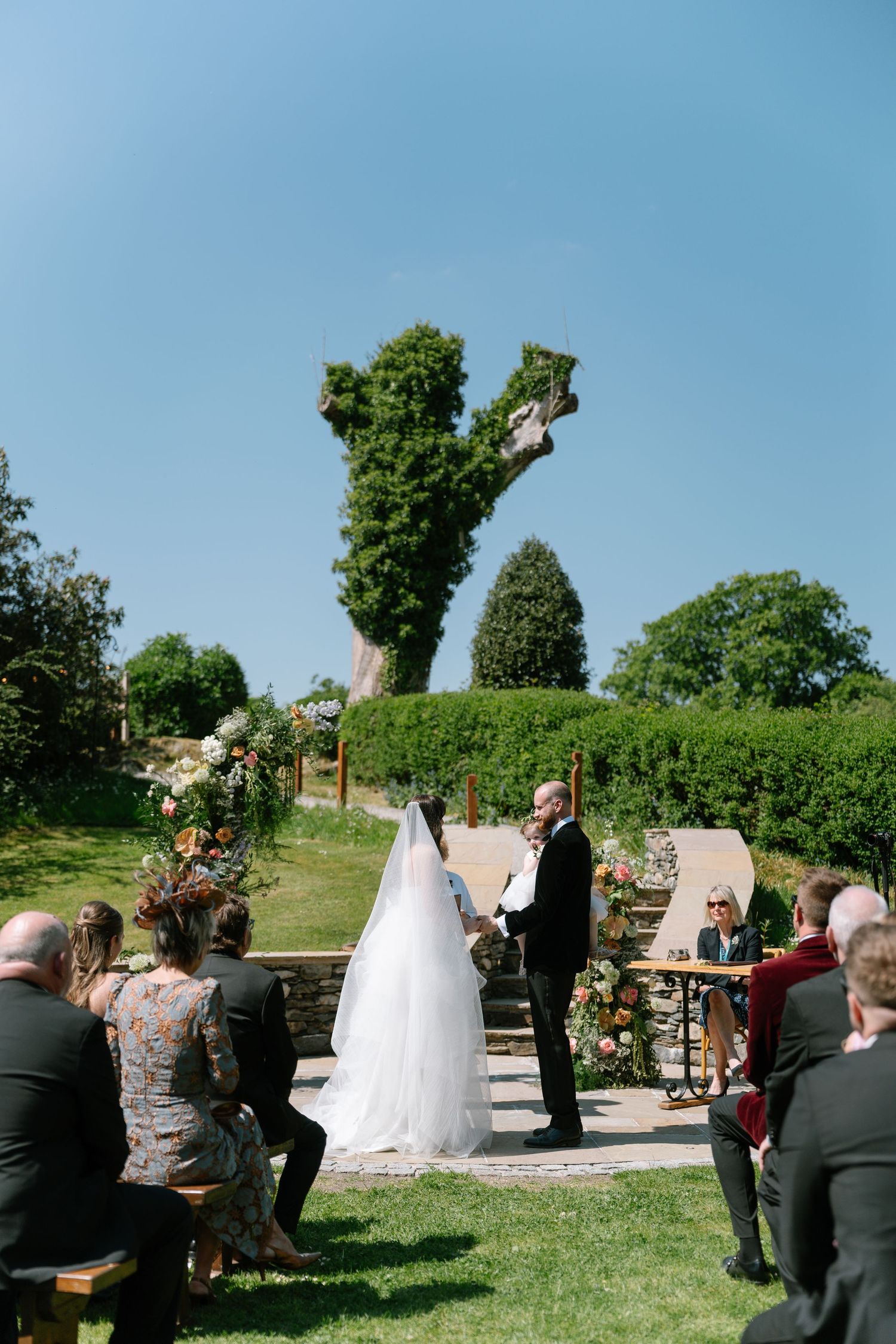 Outdoor wedding ceremony on grassy lawn with stone buildings and blue sky backdrop.