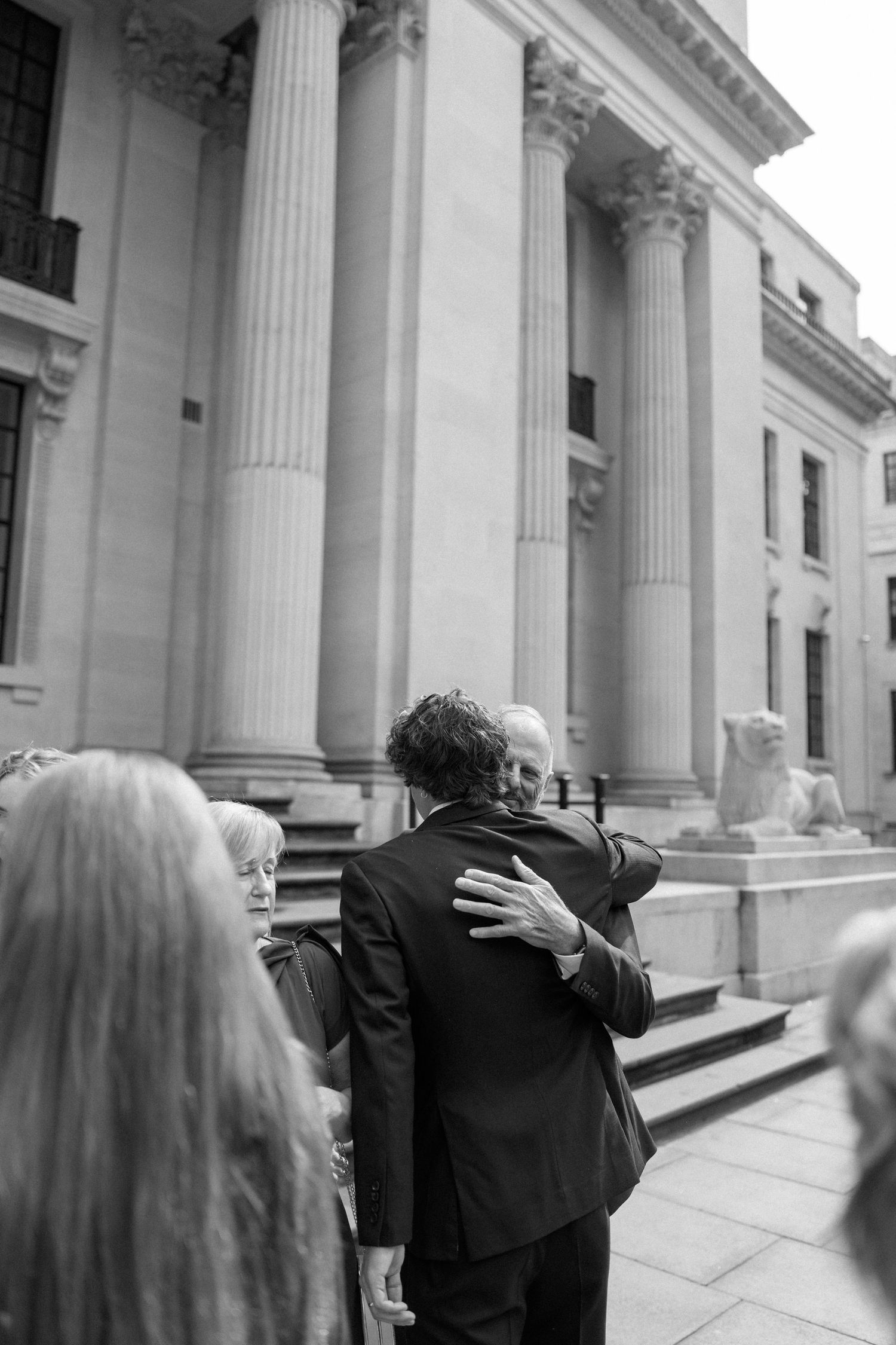 Emotional embrace between wedding guests on stone steps with classical columns.