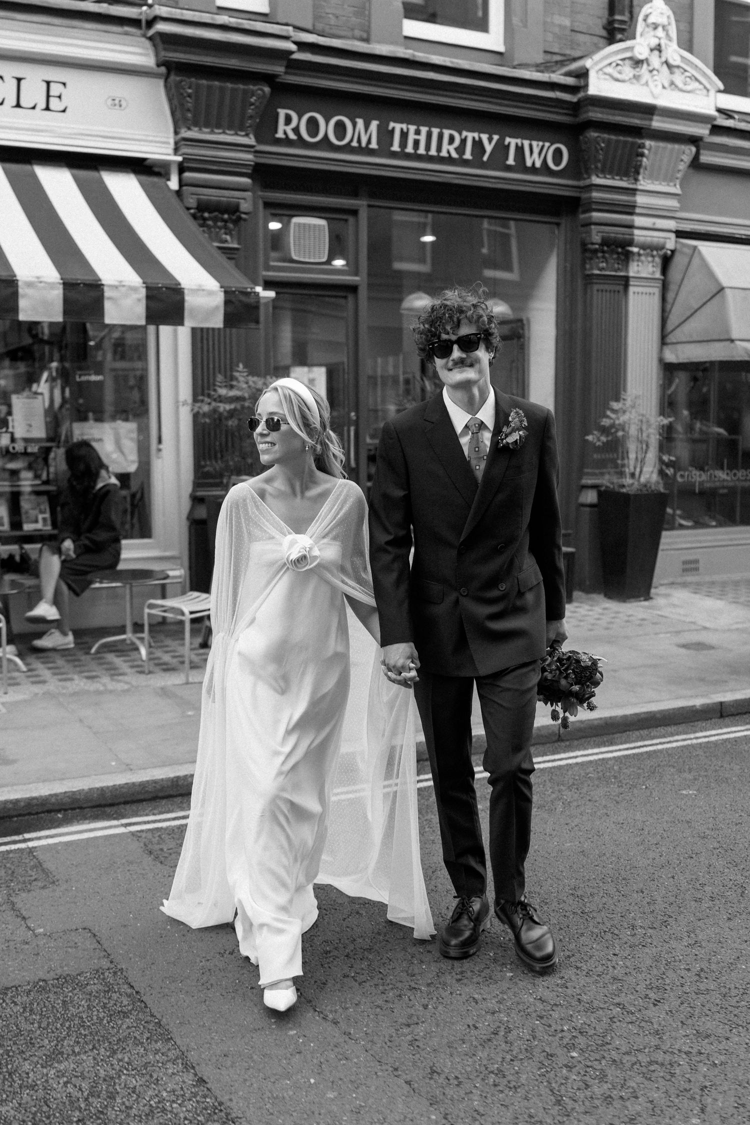 A couple walks past Room Thirty Two and Monocle storefronts in wedding attire.