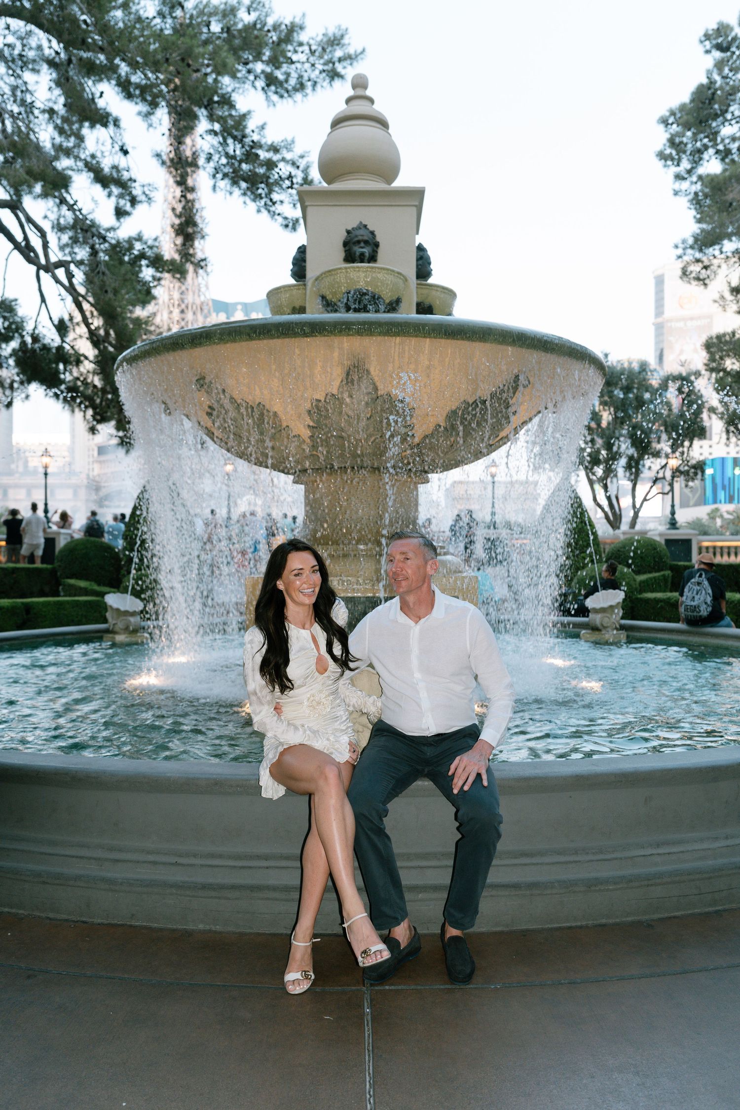 A couple sits together by a large outdoor fountain with water spraying behind them.