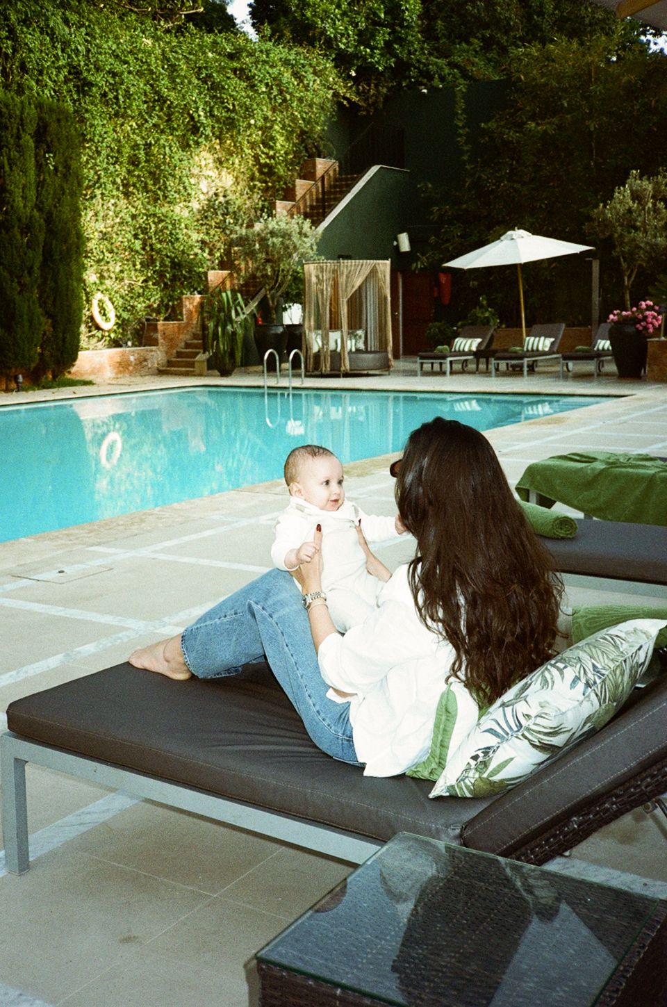 A woman with long dark hair holds a baby poolside on a lounge chair, surrounded by lush greenery.
