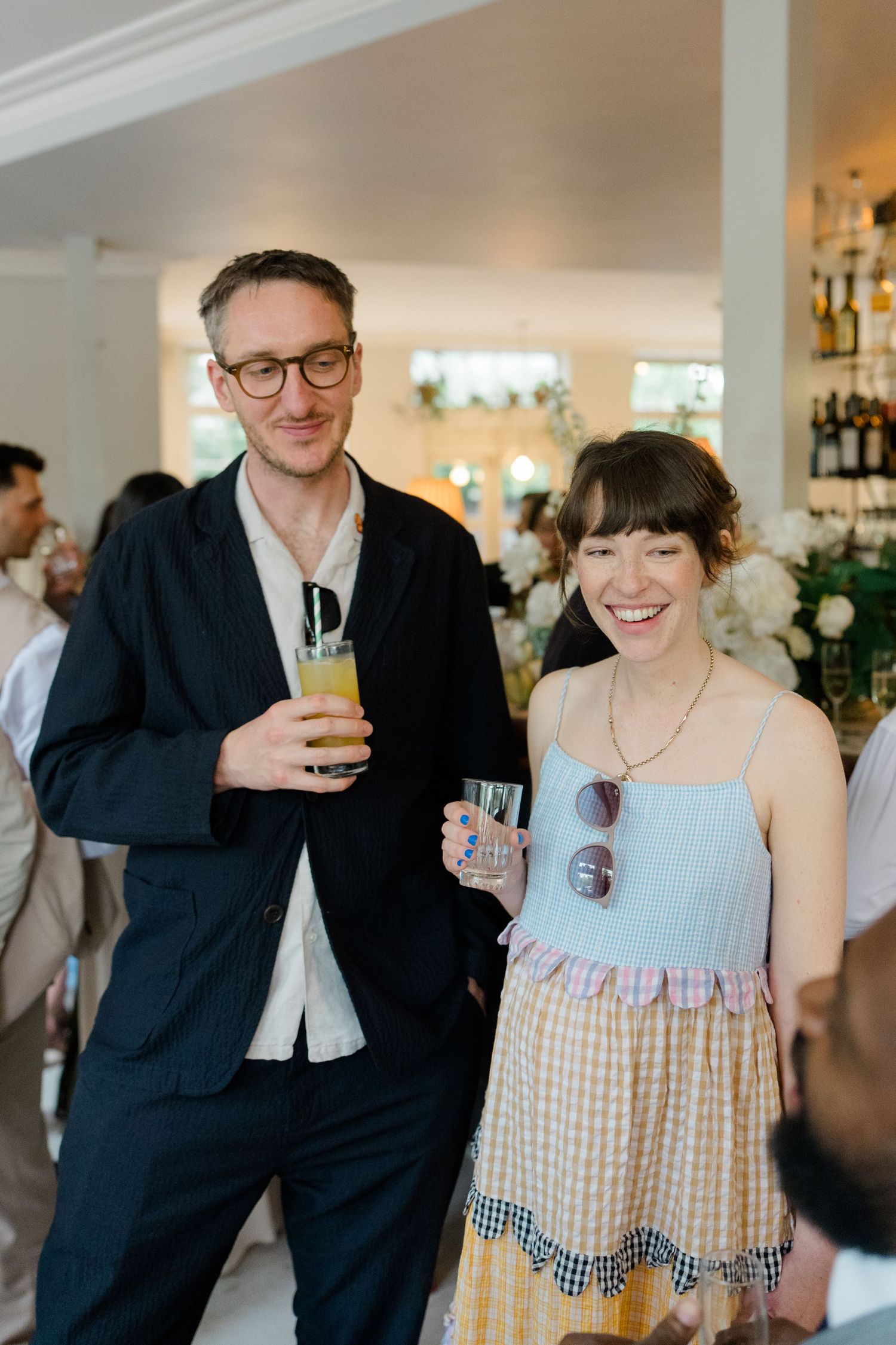 Smiling couple holding drinks pose together at an elegant wedding reception in a bright white venue.