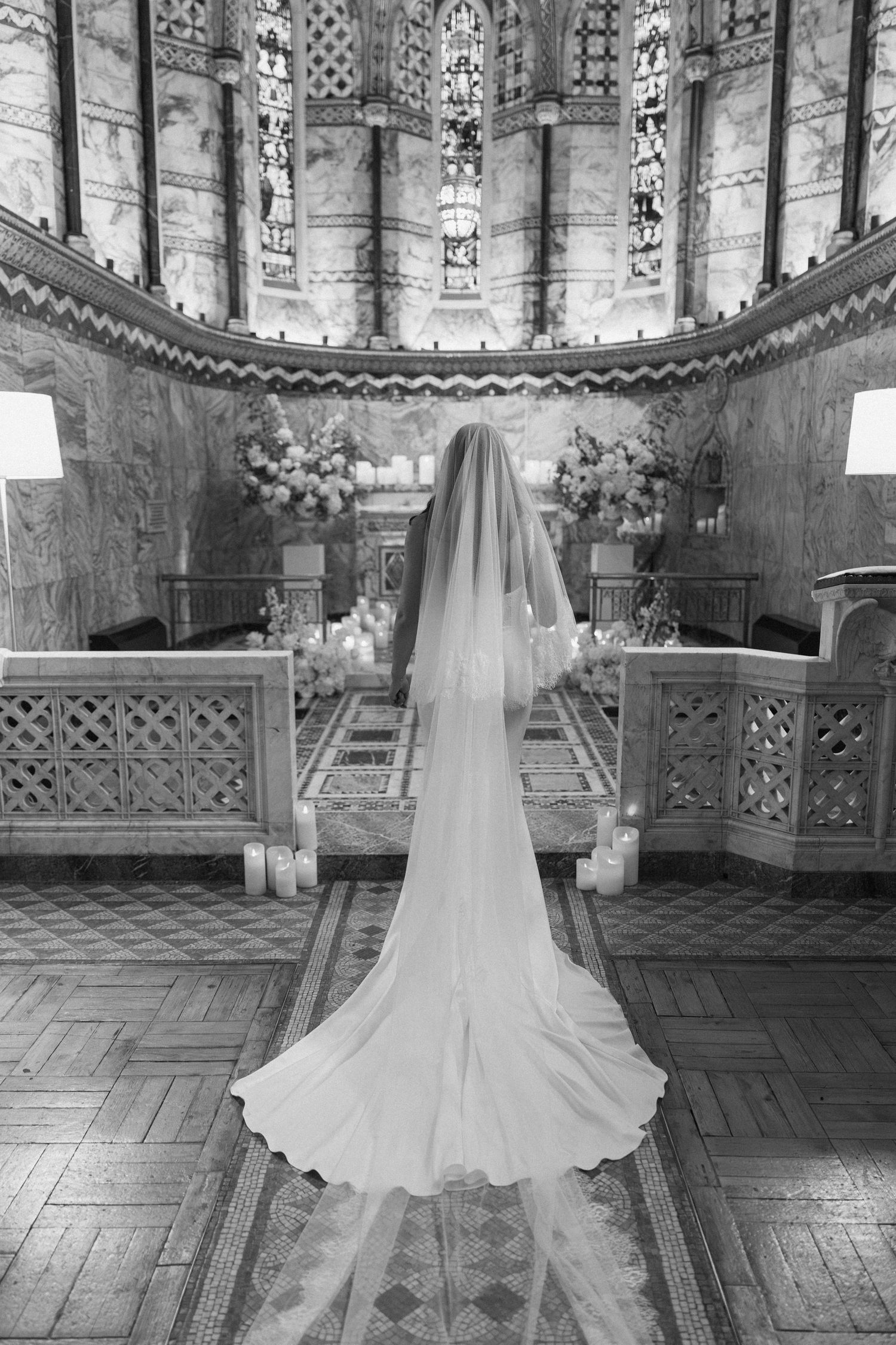 Bride in wedding gown and long veil stands at church altar with stained glass windows in black and white.