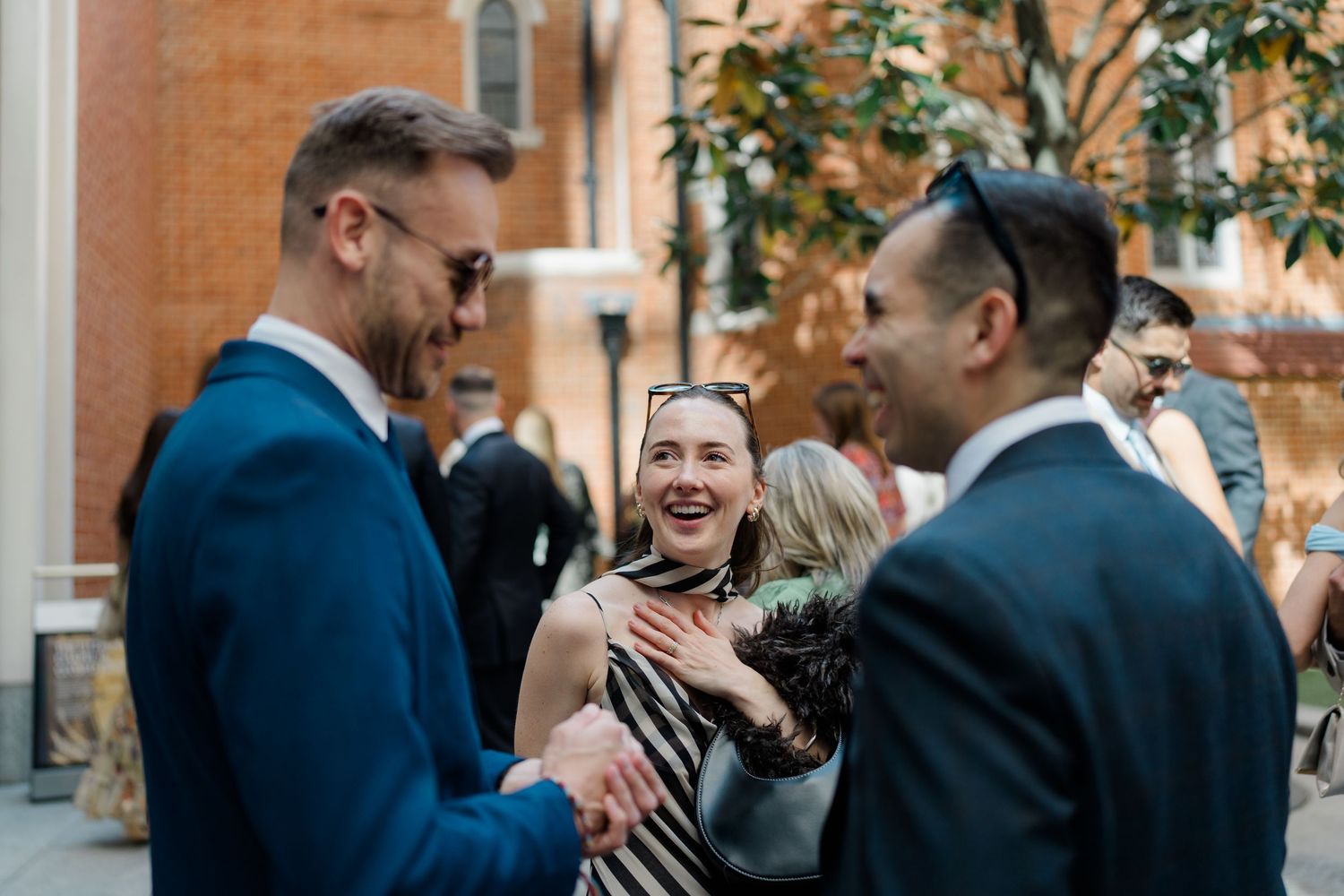 Guests laughing and socializing outdoors at a wedding reception near a brick building.