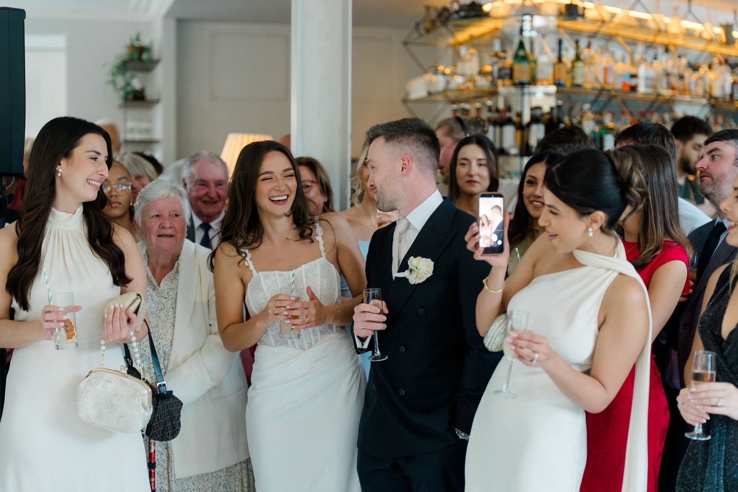 Wedding party guests laughing and holding champagne glasses during a reception celebration indoors.