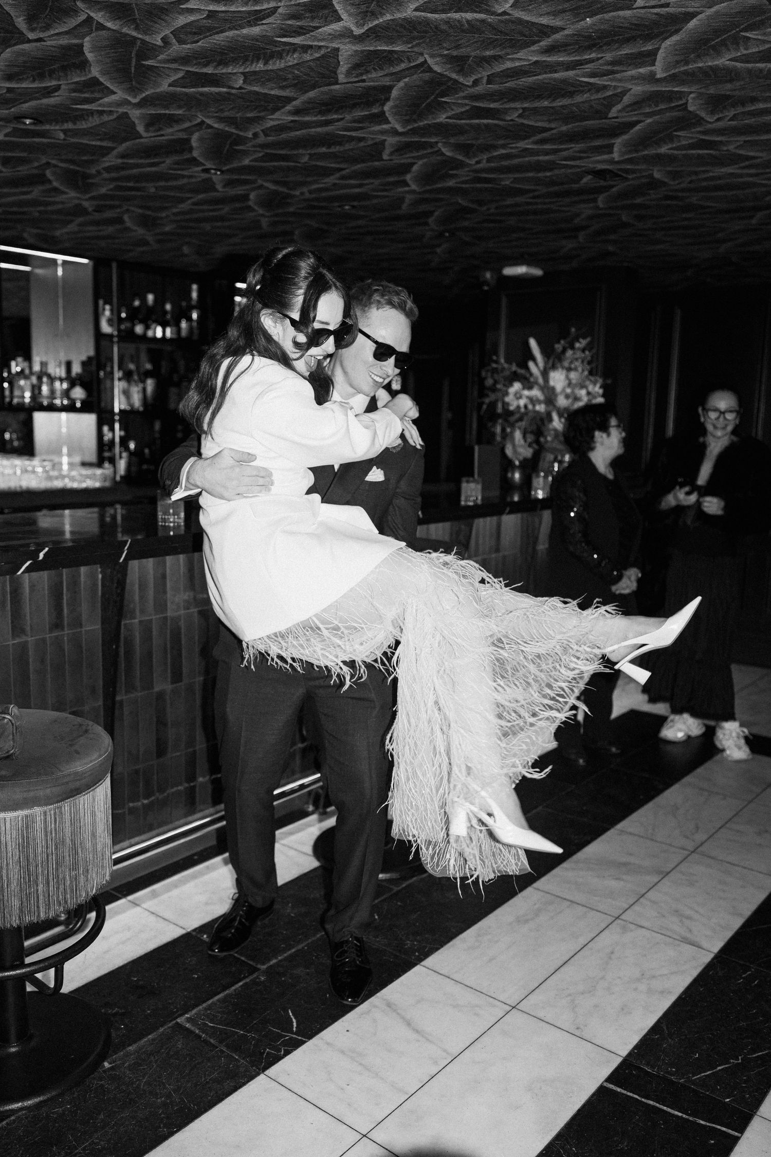 Black and white photo of groom lifting bride in feathered dress at a stylish bar venue.