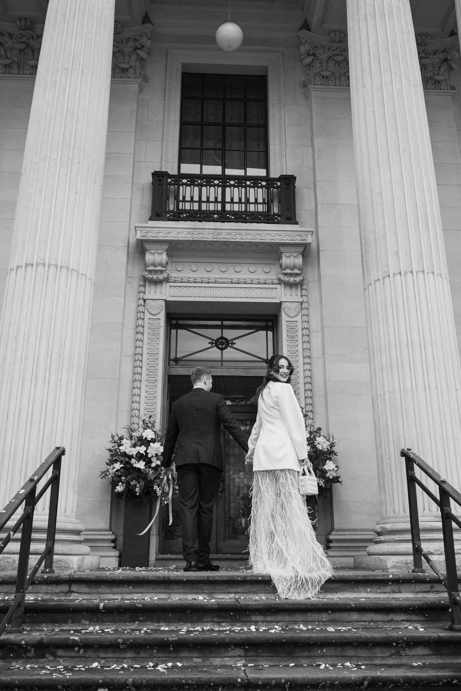 Bride in white fringe skirt and groom ascending steps of a grand neoclassical building.