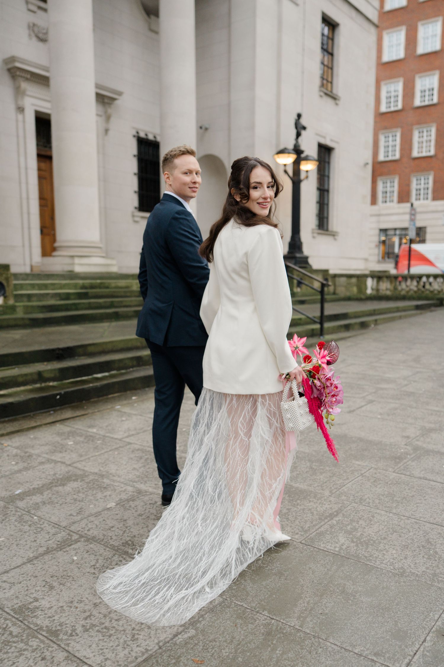 Bride in white blazer and lace feather skirt holding pink bouquet poses with groom in navy suit outside grand building.