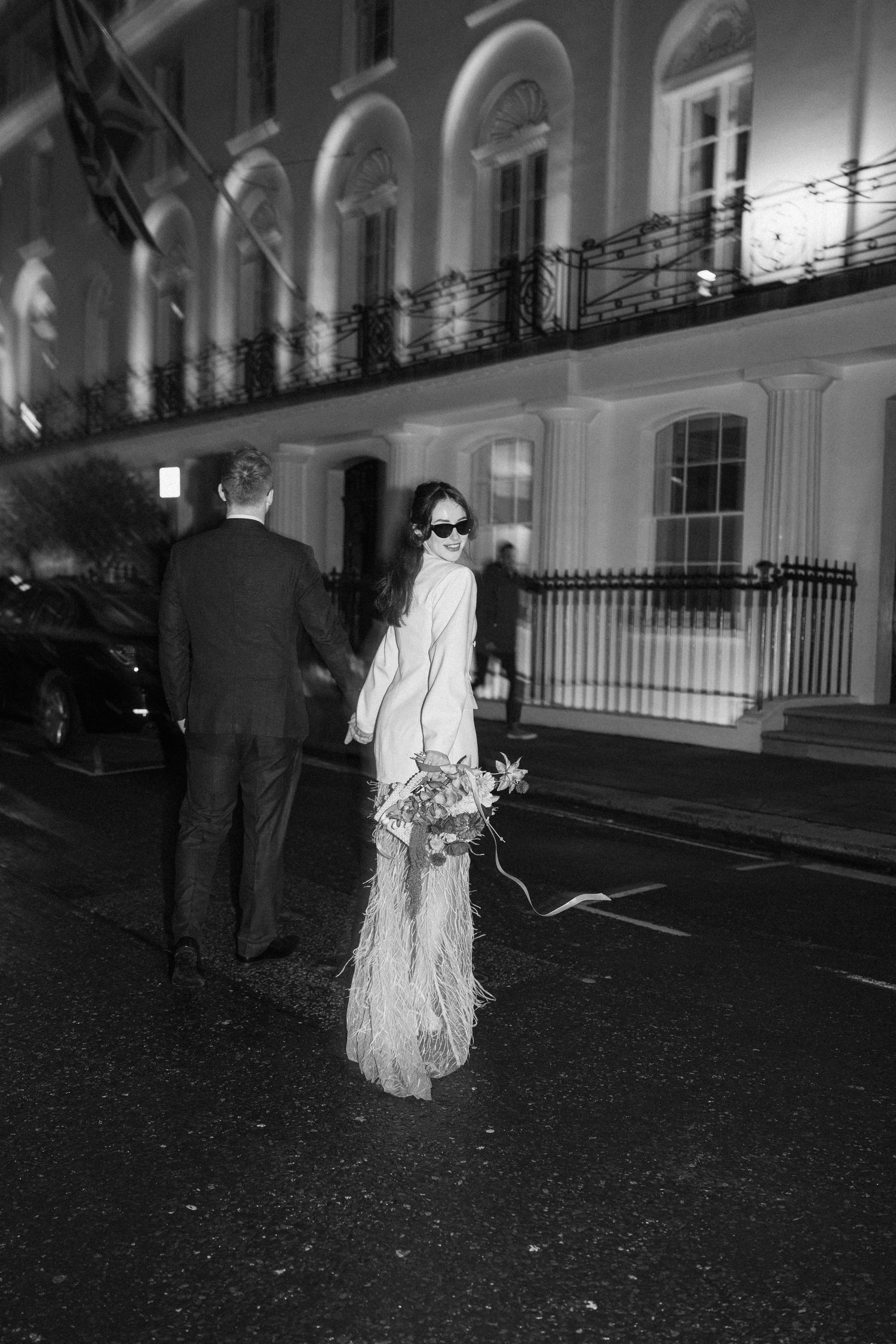 Bride in white feather skirt with bouquet and groom walk London street at night in black and white.