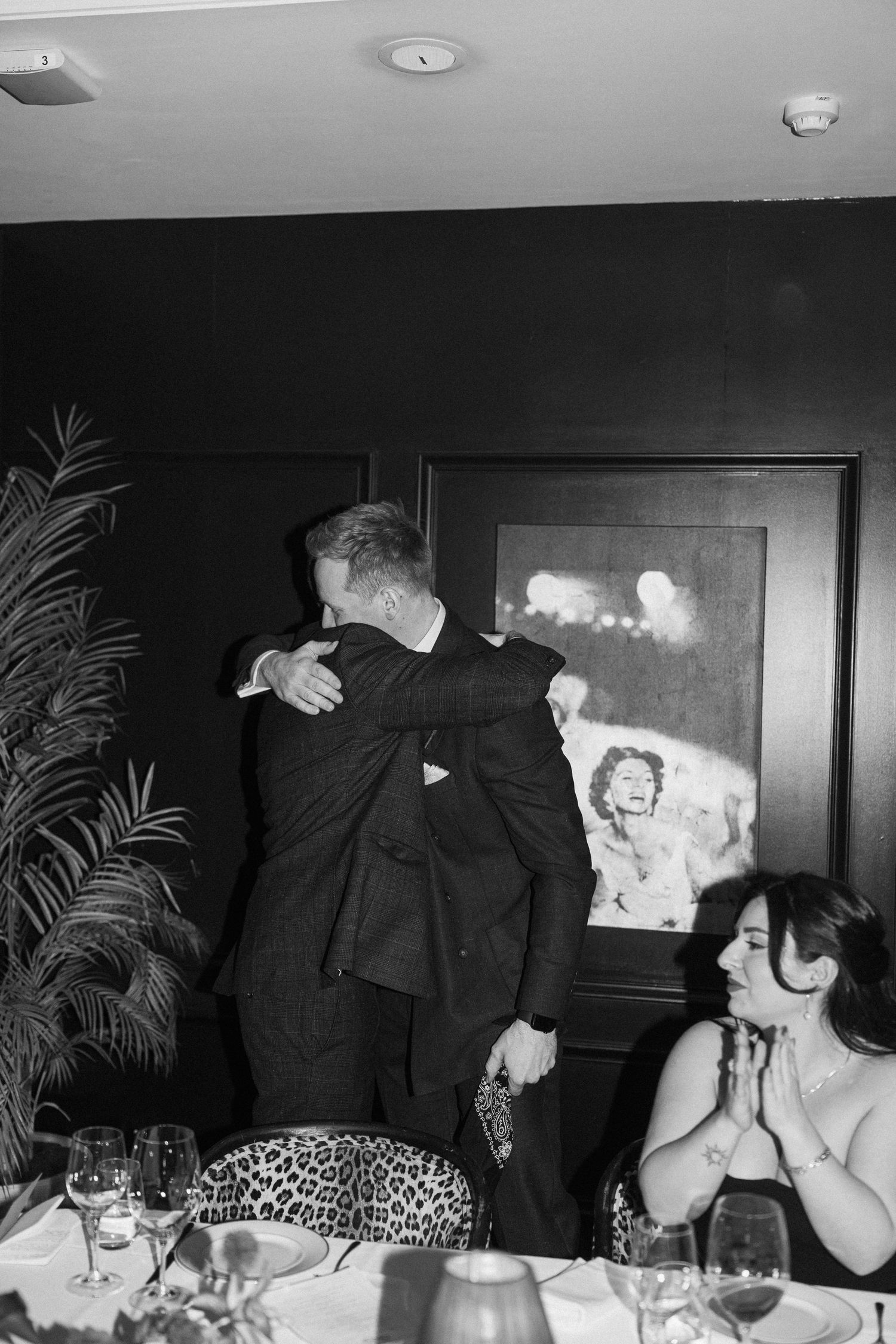 Two men embracing at an elegant dinner event in black and white, with a framed photo on the dark wall behind them.