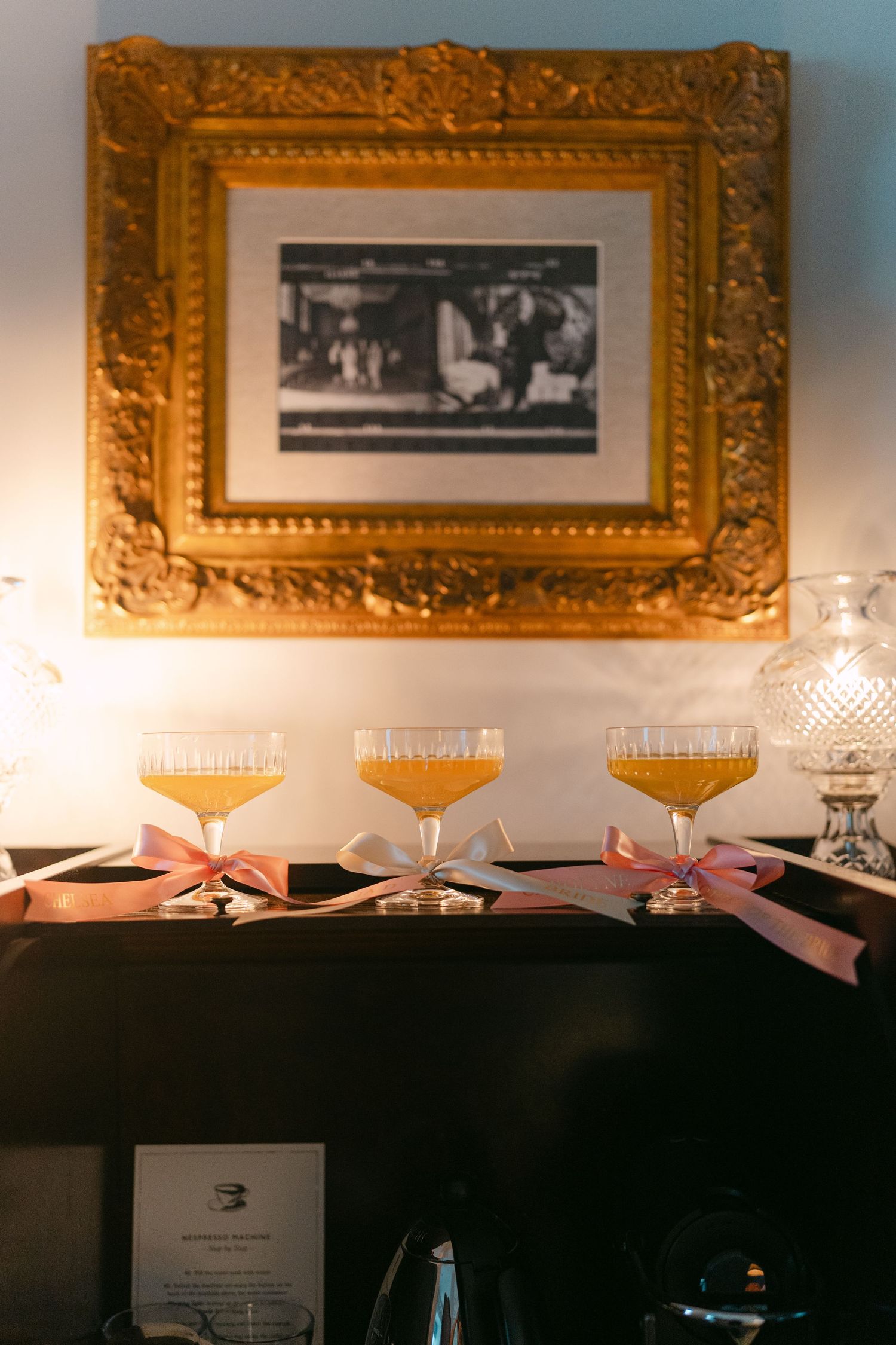 Elegant cocktail glasses filled with orange drinks displayed in front of vintage framed photographs on a wall.