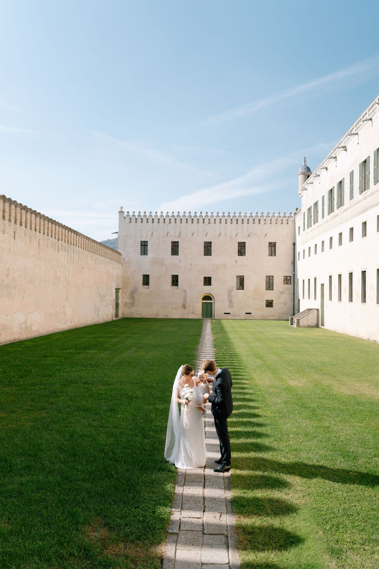 Wide landscape view of a wedding ceremony against dramatic white castle walls and green lawn.