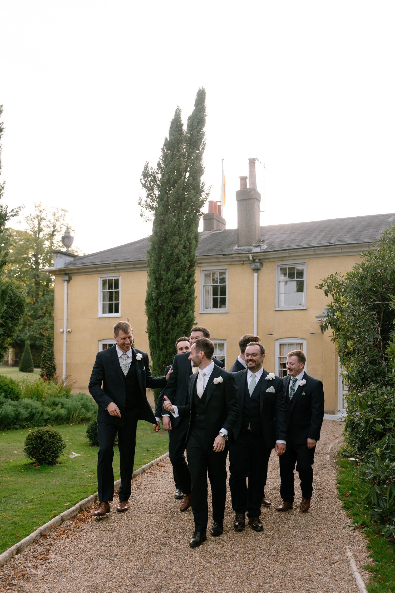 Groomsmen in black suits walk together along a gravel path outside a yellow house.
