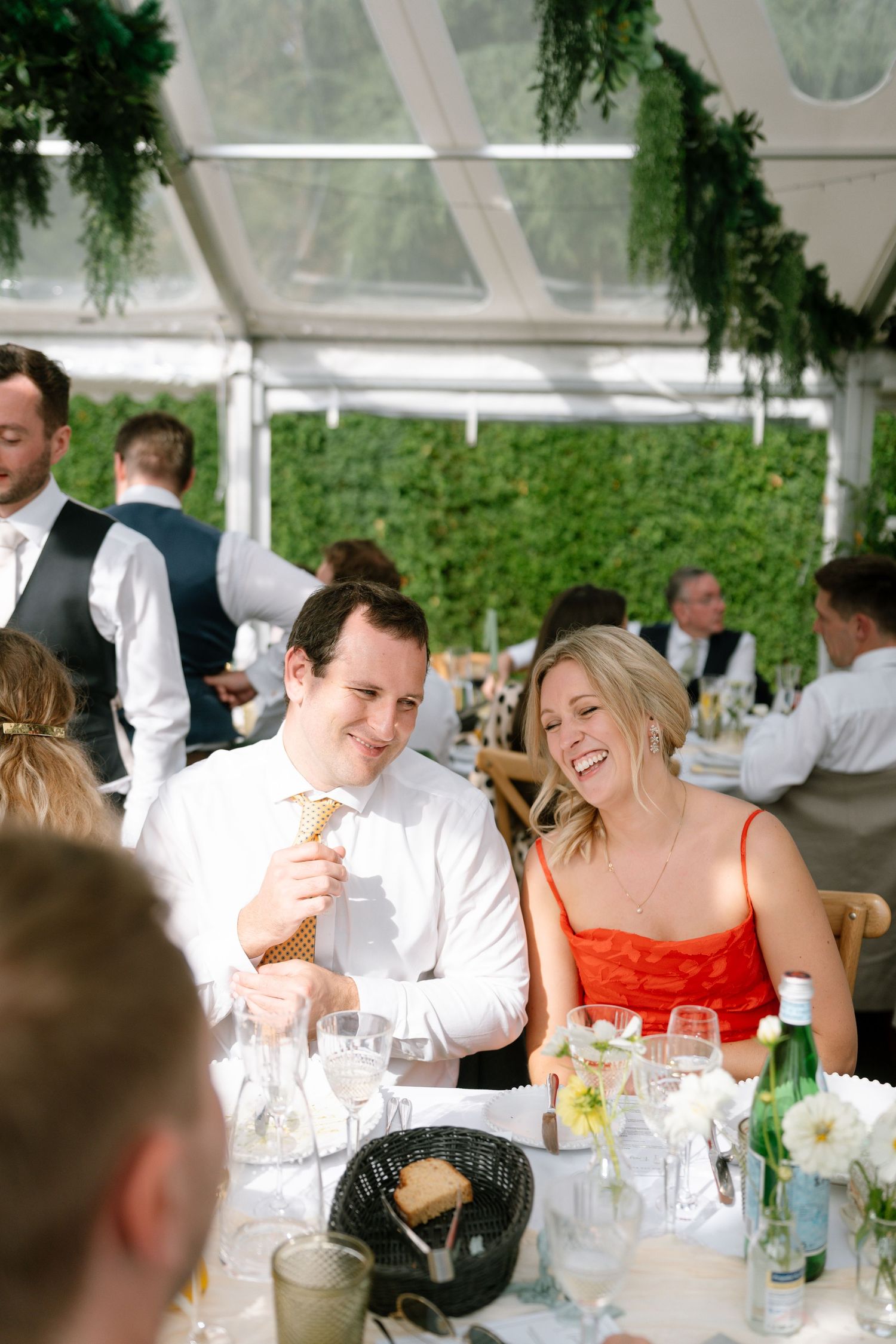 Guests enjoying a celebration dinner under a clear tent with hanging greenery.