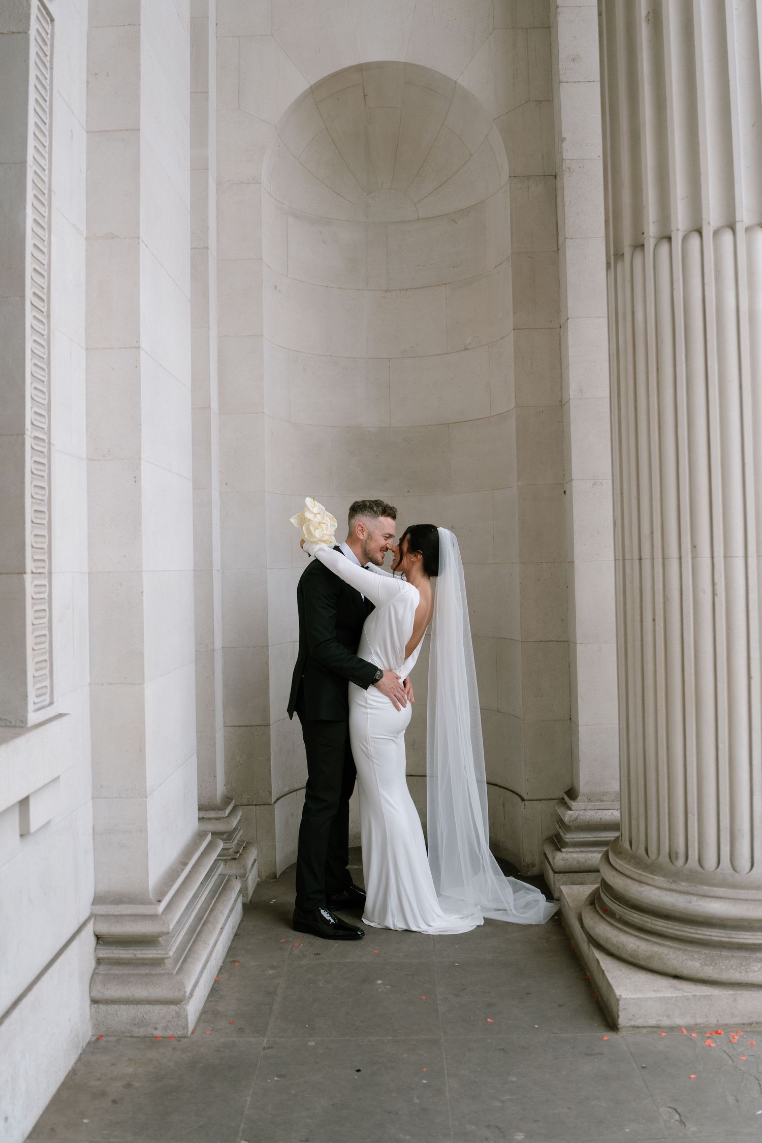 Wedding couple posing between classical stone columns in elegant architectural setting.