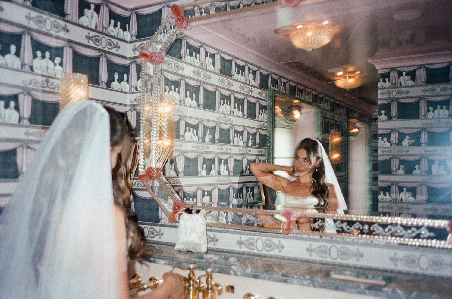 Bride in white veil adjusts hair while reflected in ornate mirrored wall with decorative chandelier details.