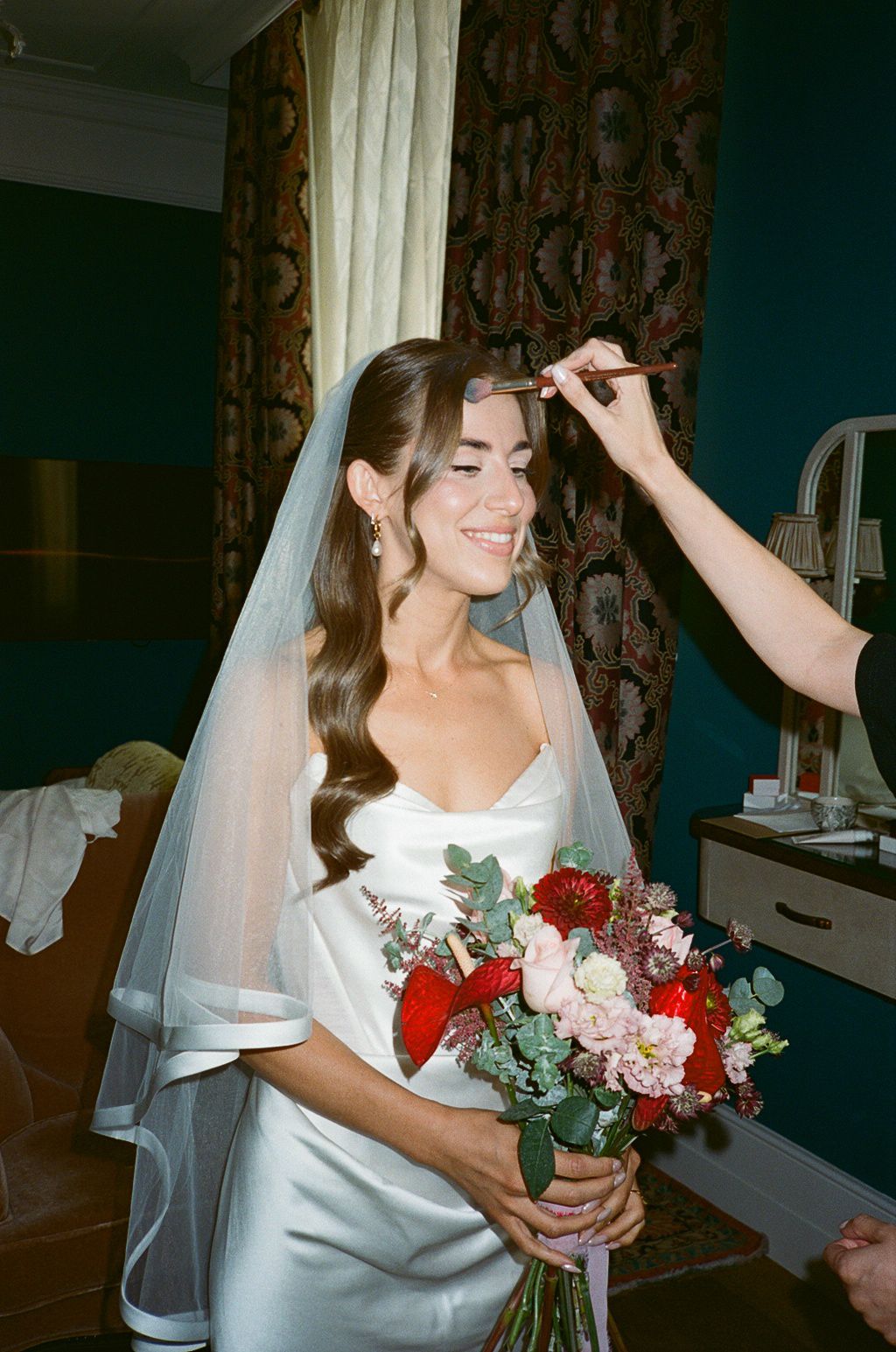 Smiling bride in white satin gown and veil holds red bouquet while makeup artist applies finishing touches.