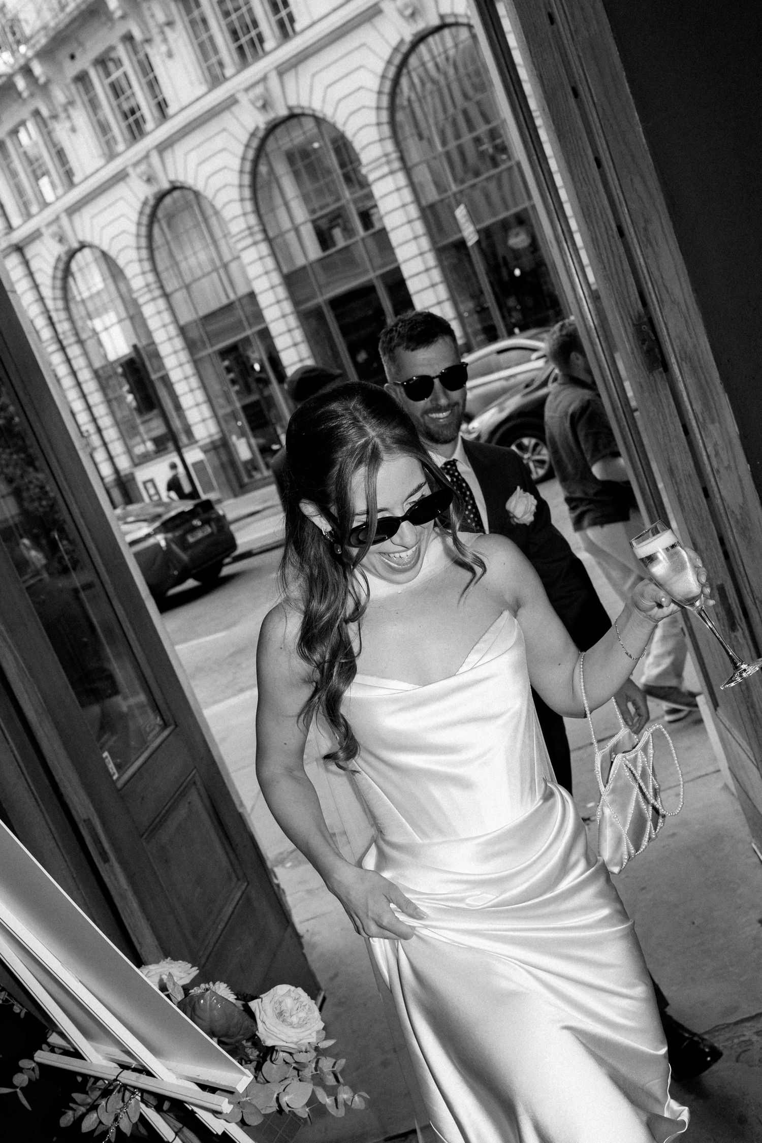 Black and white photo of bride in sunglasses holding champagne exiting elegant building.