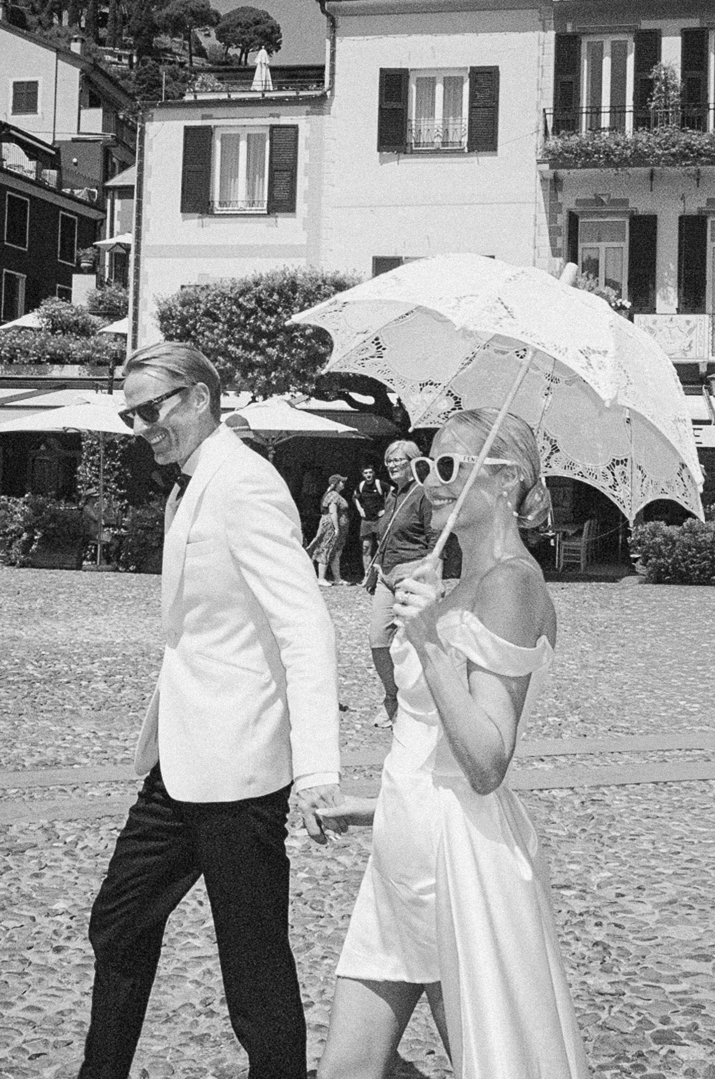 Black and white photo of newlyweds walking through Italian piazza, bride holding decorative white parasol.