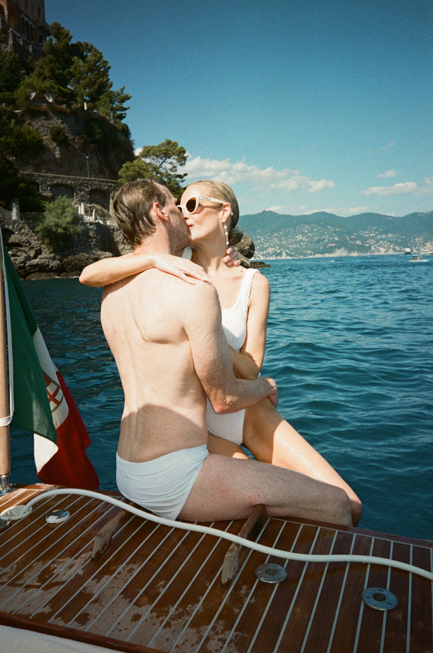 Couple embracing and kissing on wooden boat with Italian flag and coastal cliffs behind.