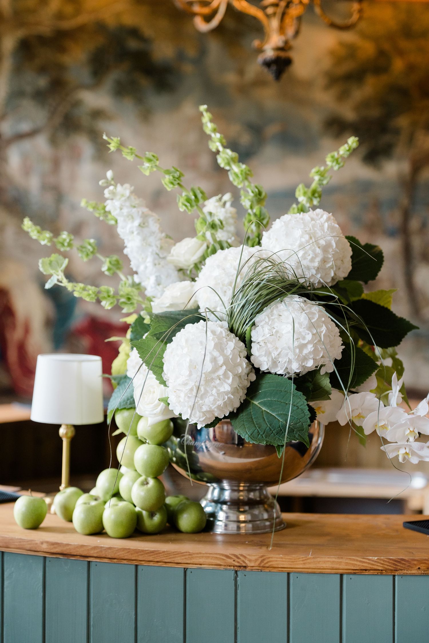 White hydrangea and delphinium floral arrangement with green apples in silver bowl on wooden table.