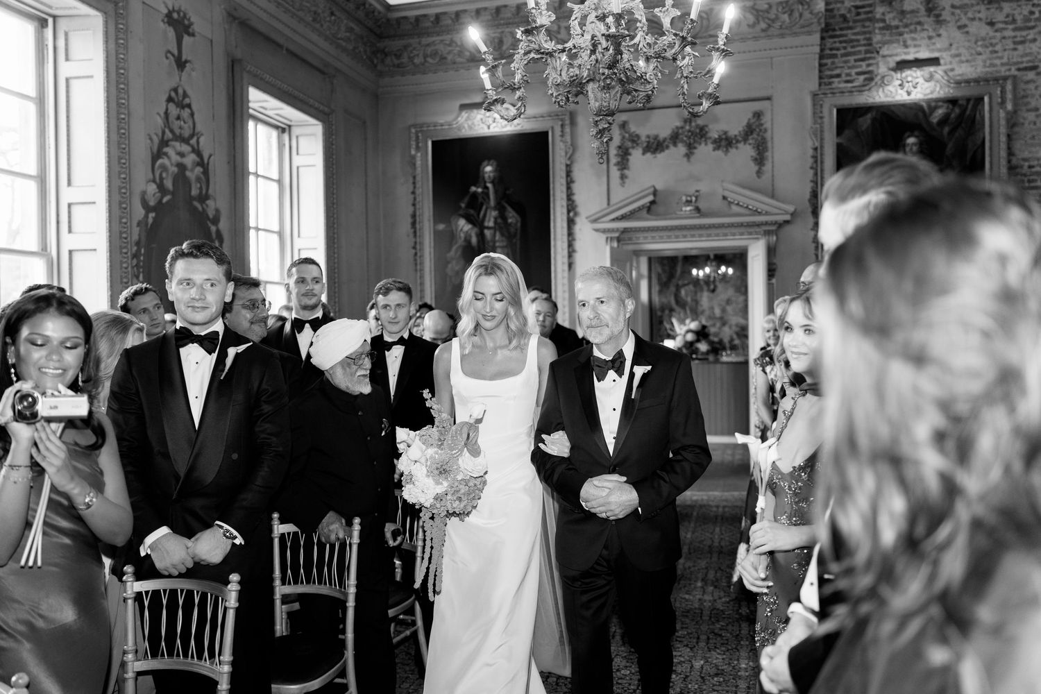 Bride walks down the aisle with her father in a grand historic wedding venue, surrounded by guests in formal attire.