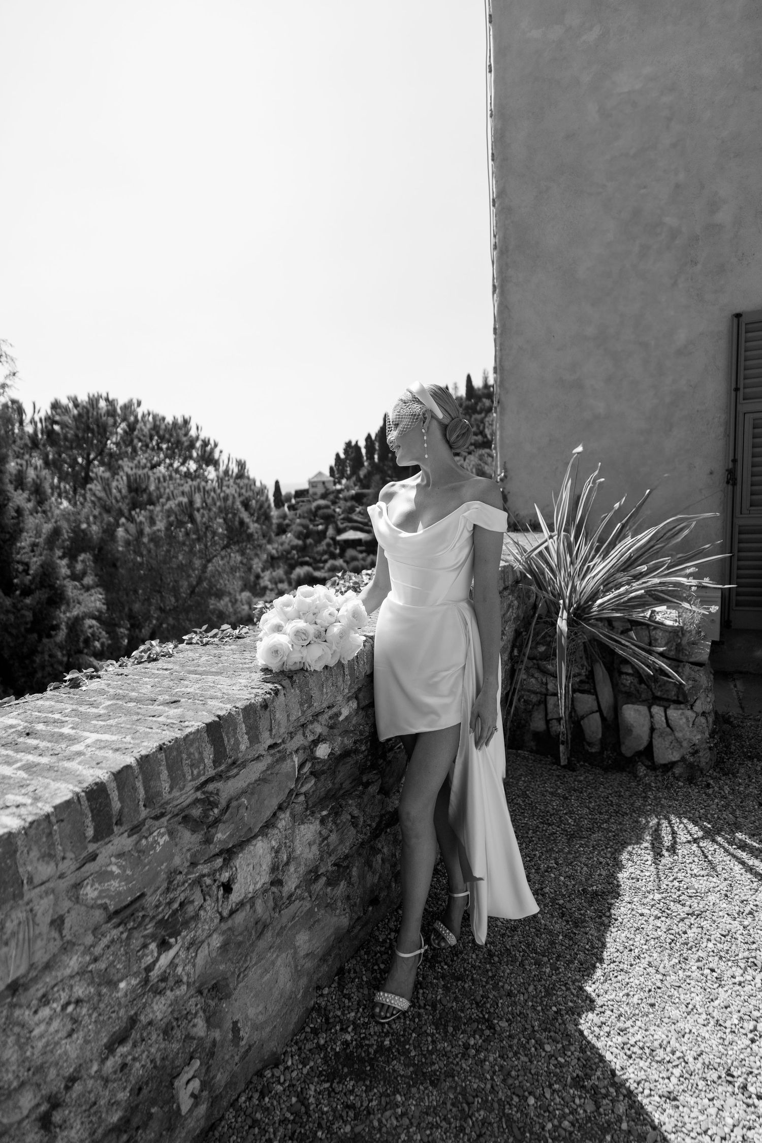 Black and white photo of bride in short wedding dress leaning against stone wall with bouquet.