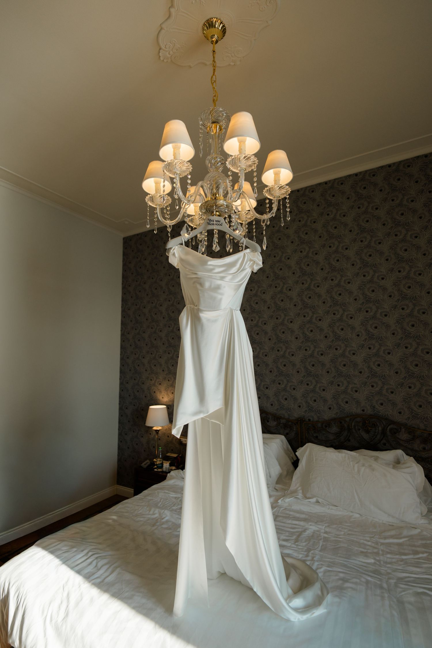 Elegant white wedding dress hanging from a crystal chandelier above a bed in a luxurious hotel room.