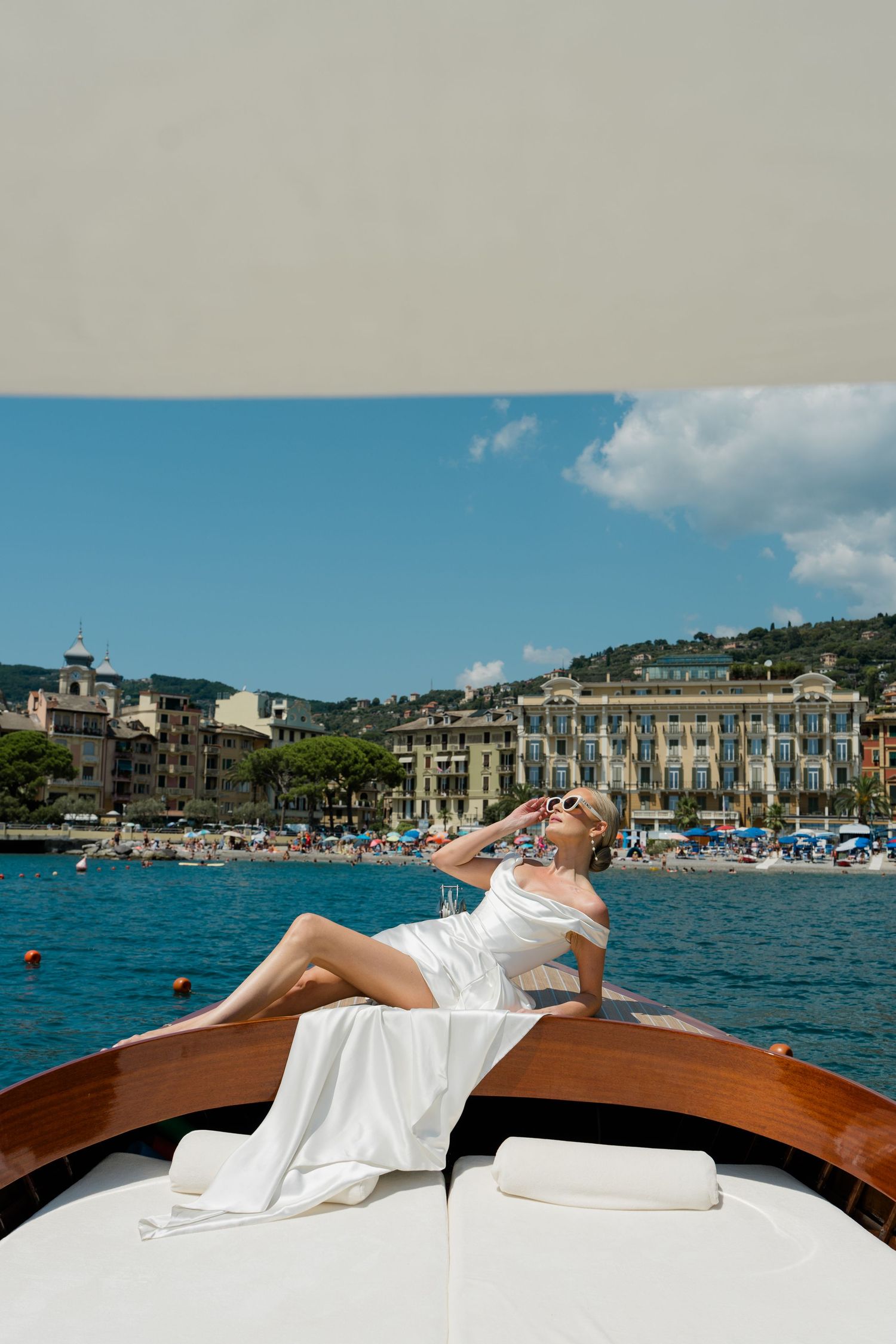 Beautiful bride in white wedding dress reclining on a luxury wooden boat with colorful Italian coastal town backdrop.