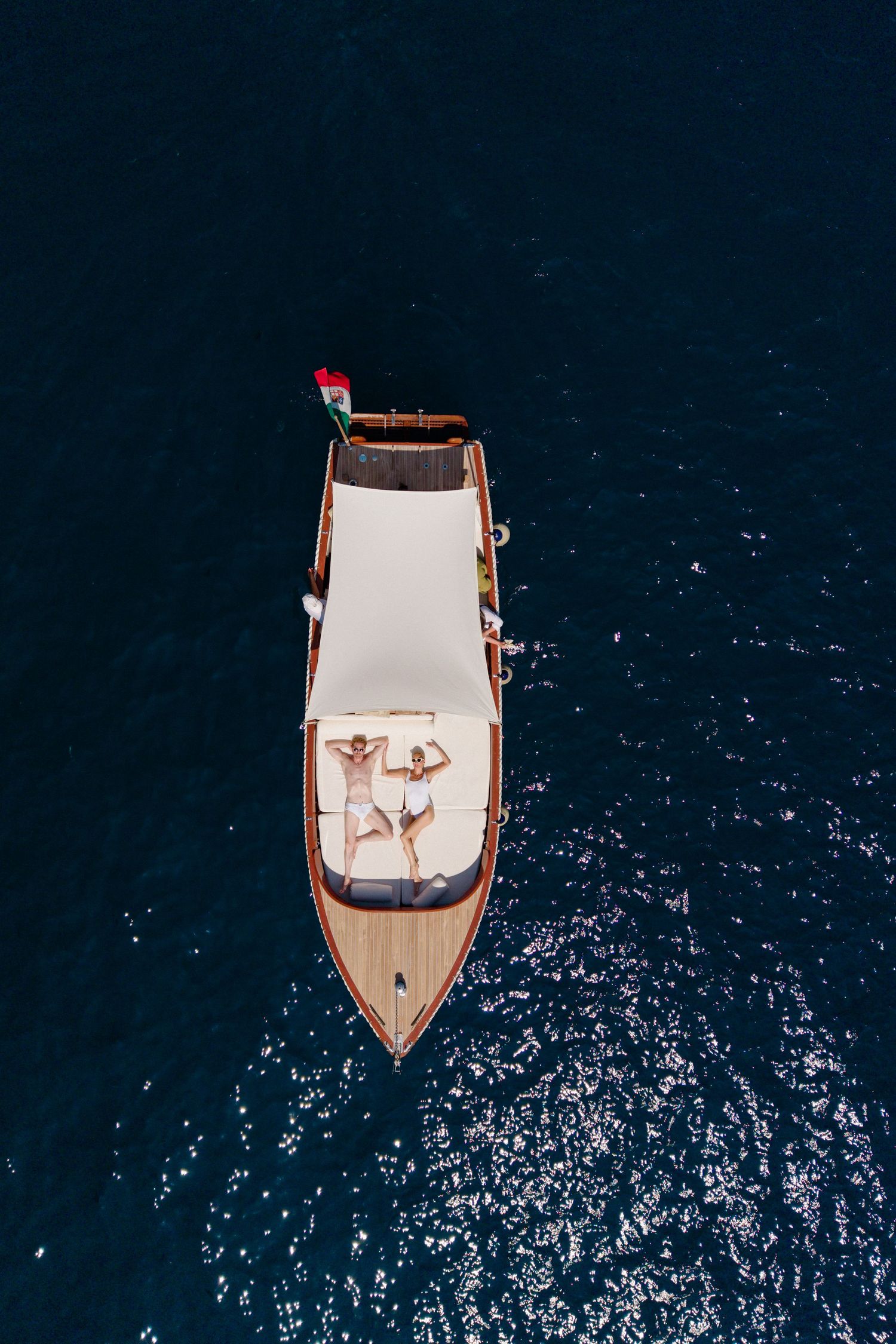 Aerial view of couple sunbathing on the deck of a wooden boat floating on deep blue water.