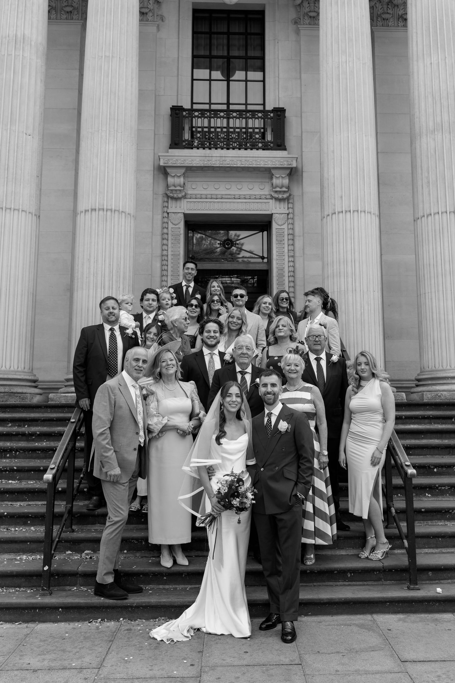 Wedding party group photo on grand stone steps with tall columns at a classic venue in black and white.