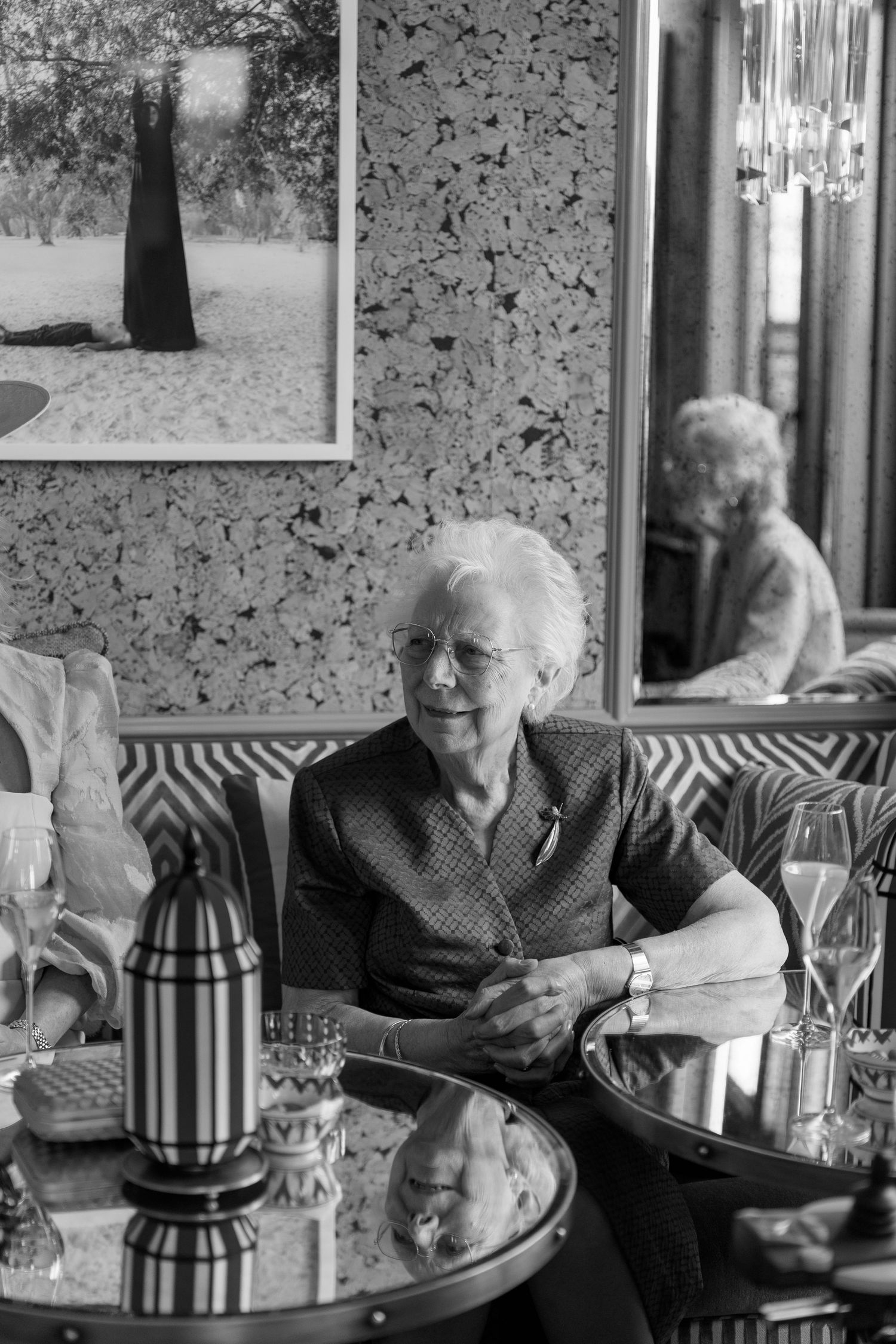 Elegant elderly woman sitting at a restaurant table with champagne glasses in black and white photography.