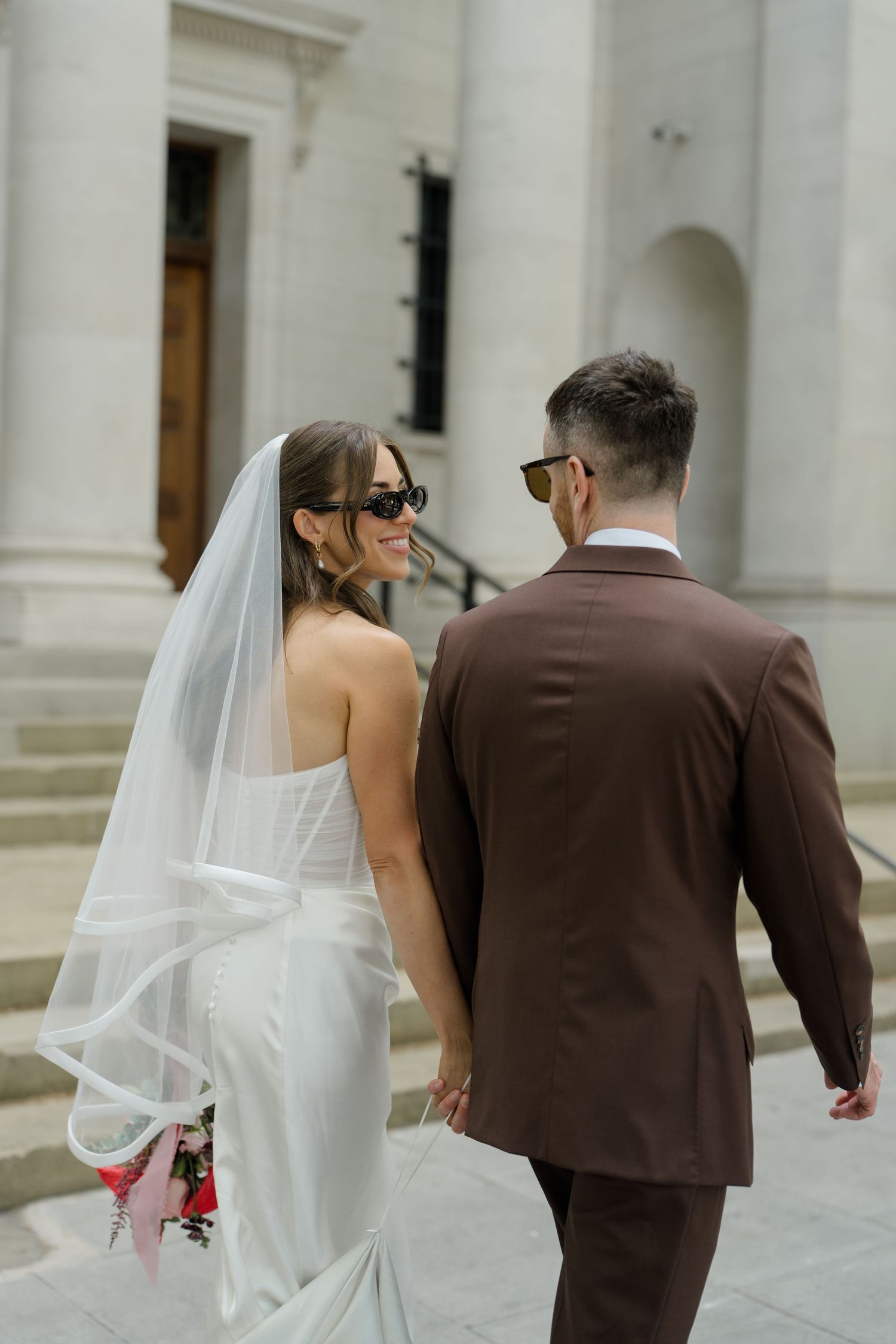 Bride in veil and sunglasses glancing back at groom in brown suit while walking up stone steps of elegant building.