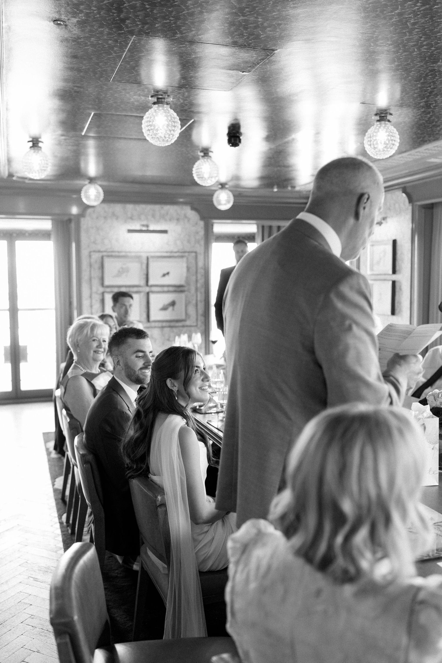 Black and white image of bride and groom seated at wedding reception table during guest speech.