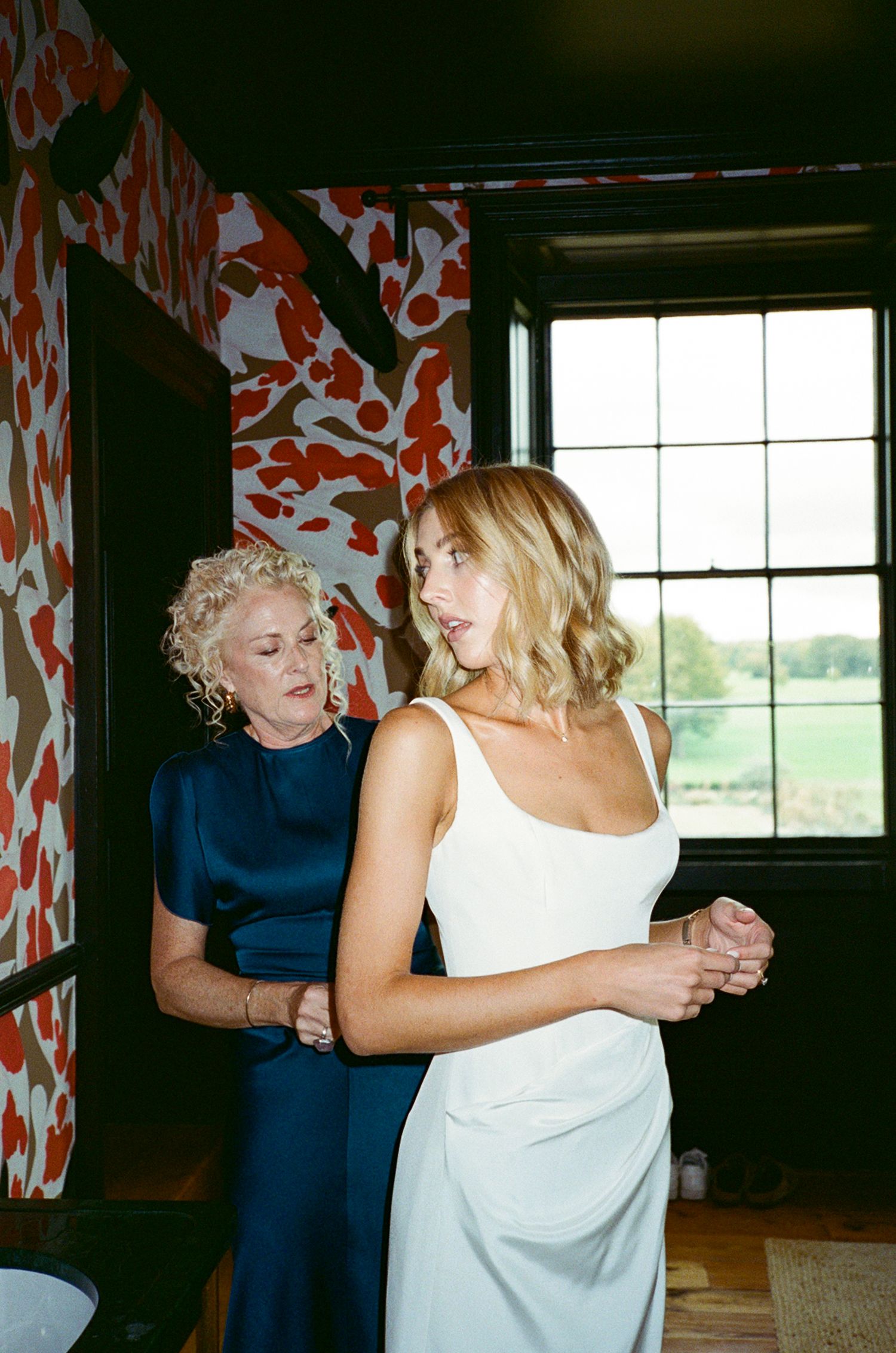 Bride in white gown being helped by older woman in navy dress near window with floral wallpaper.