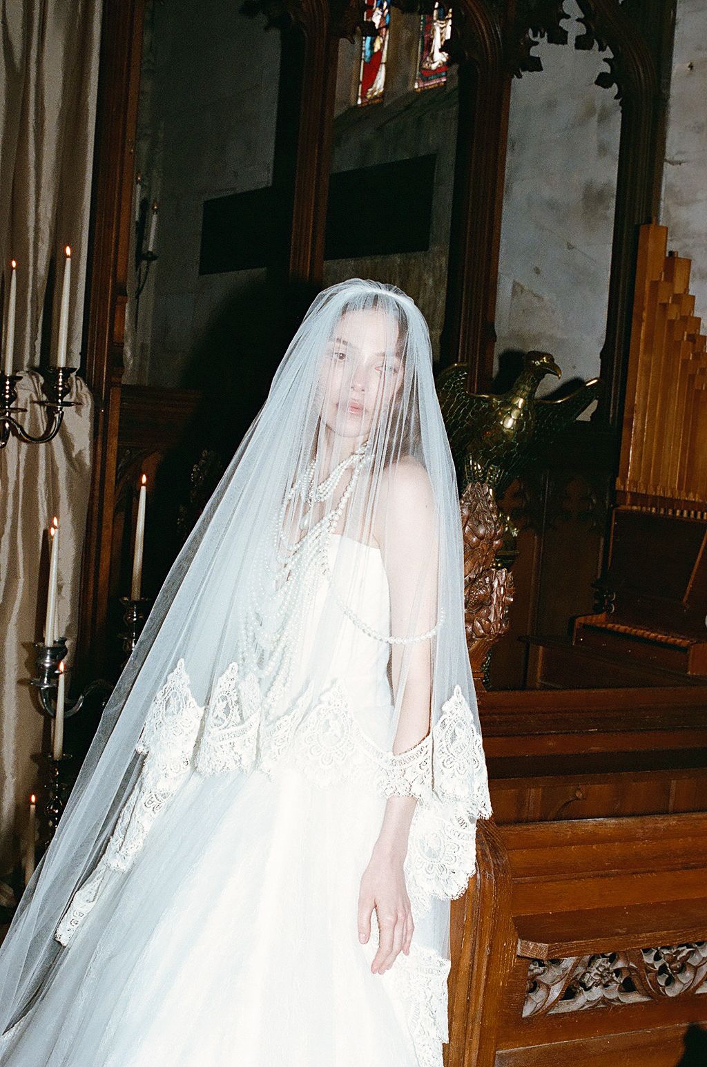A bride in a flowing white wedding gown and veil poses in a church with wooden pews and candlelit ambiance.
