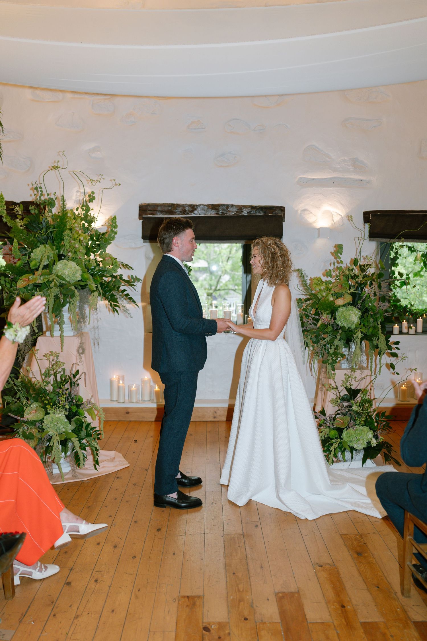 Wedding ceremony in a bright white room with wooden floors and lush greenery decorations.