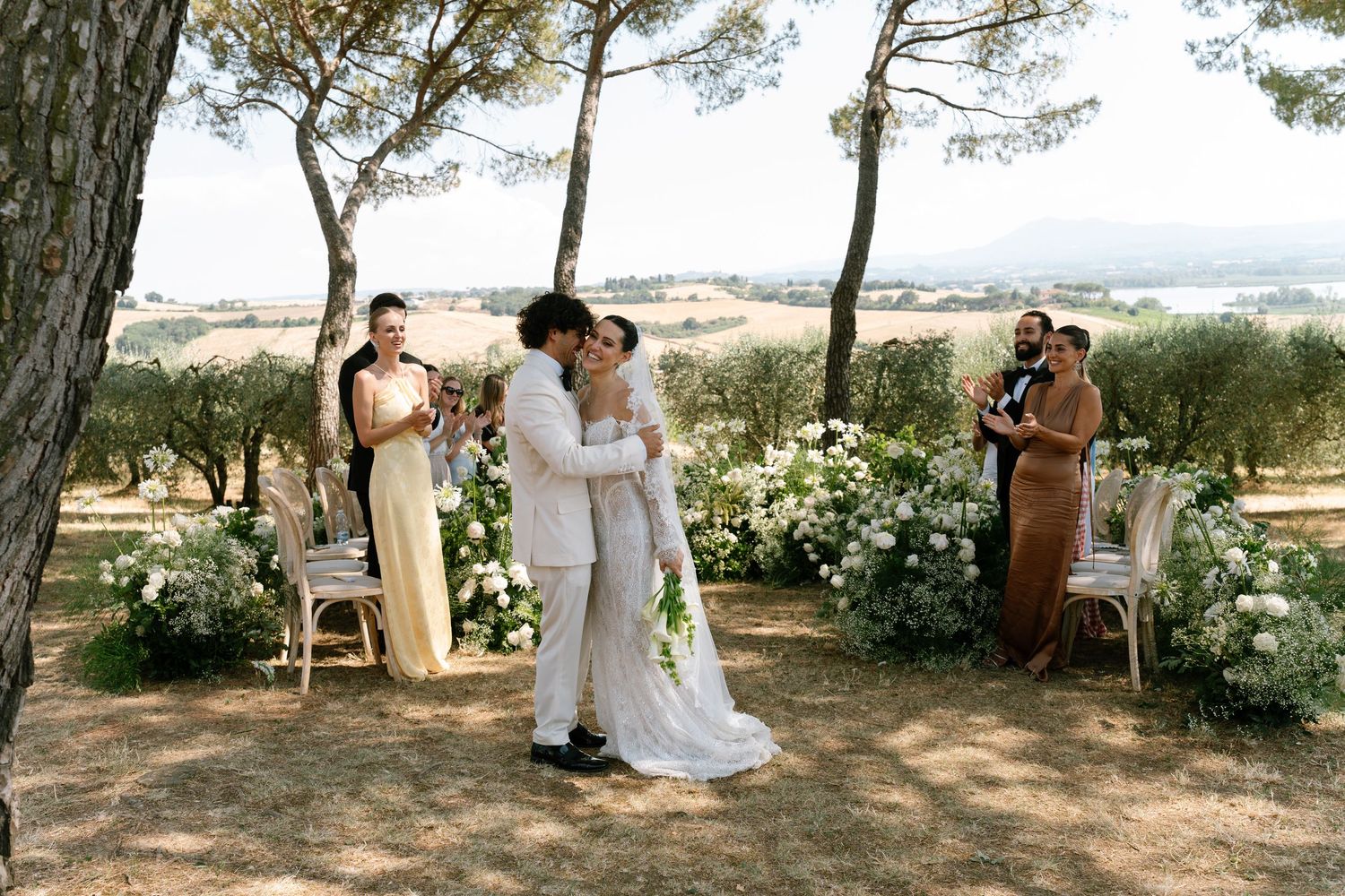 Scenic outdoor wedding ceremony setup under pine trees with white chairs and floral decorations.