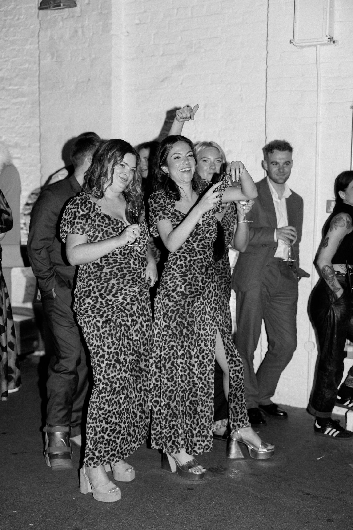 Two women in matching leopard print dresses laugh and dance together at a party in black and white photography.