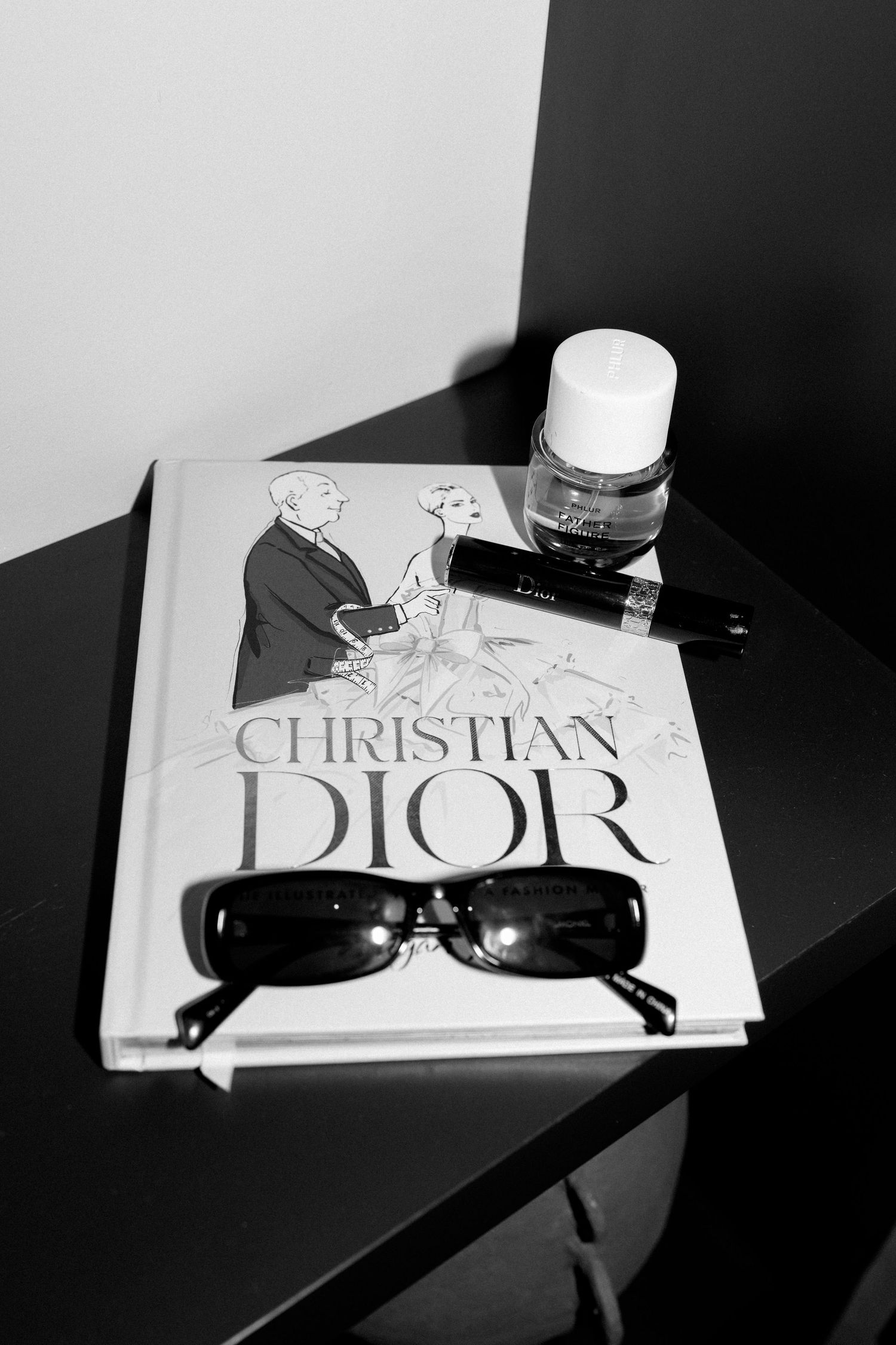 Black and white still life with Christian Dior book, sunglasses, and beauty product on dark surface.