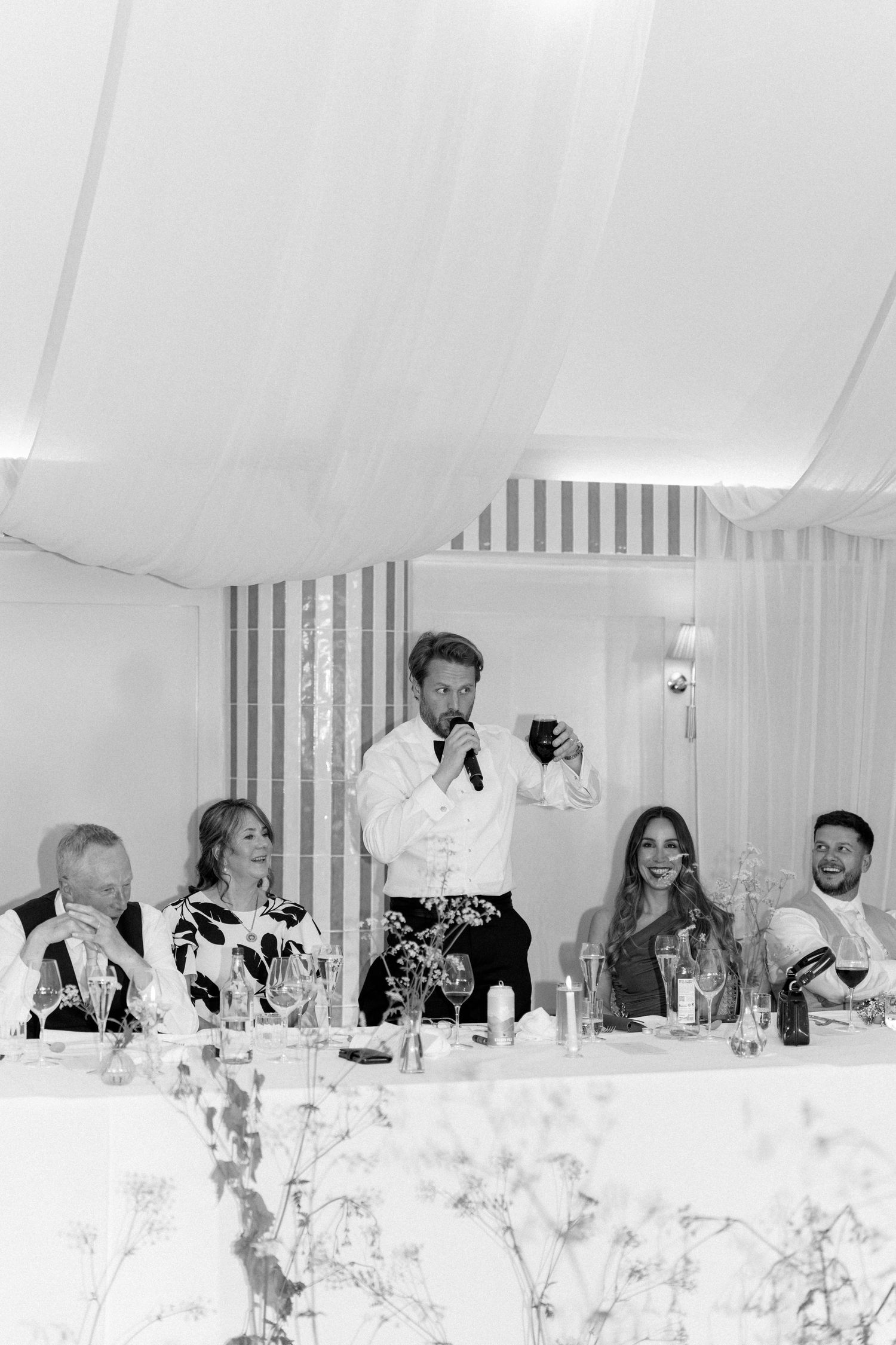 Black and white photo of best man giving speech at wedding reception while guests laugh at head table.