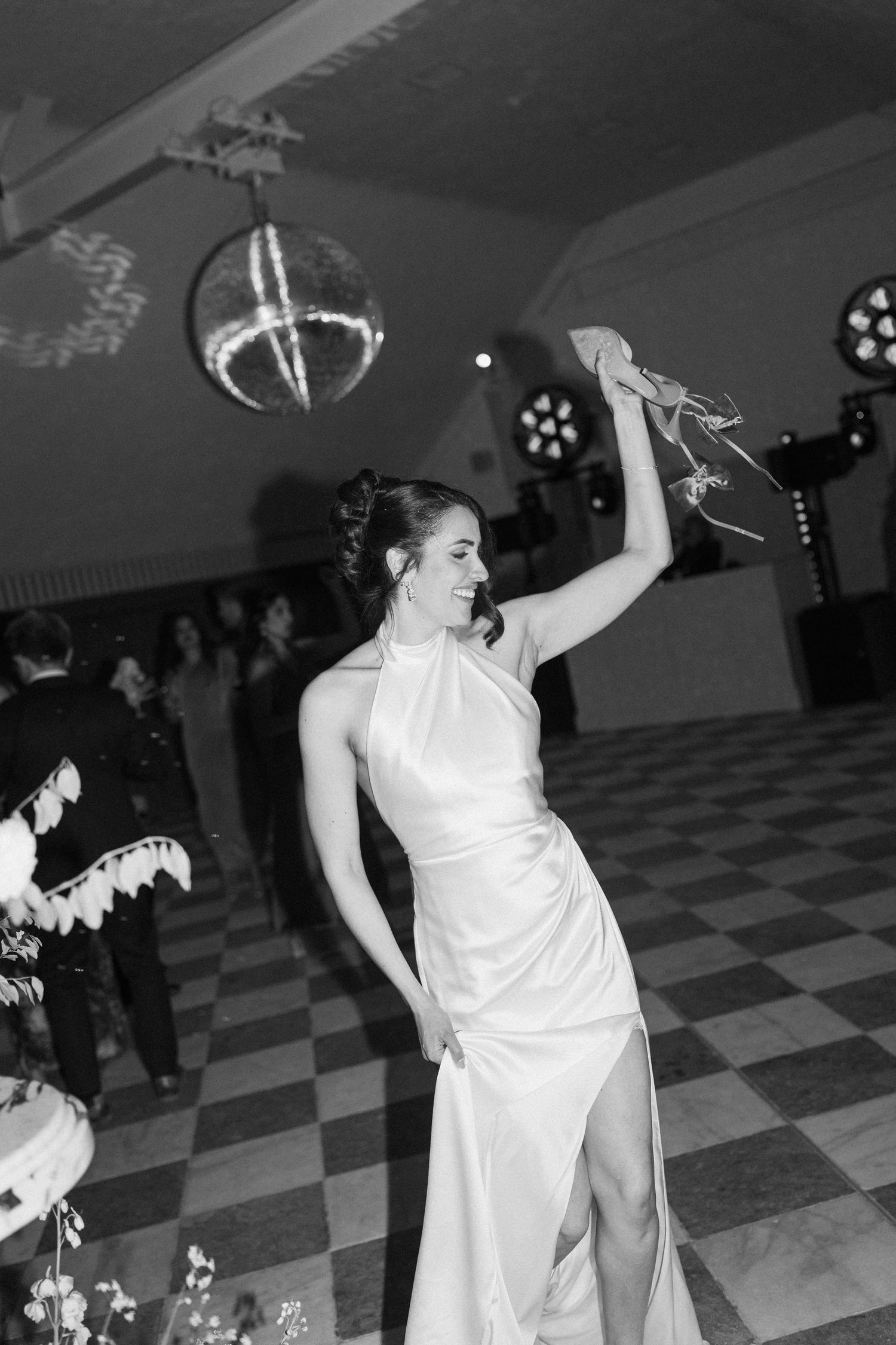 Bride dances barefoot holding her heels aloft under a disco ball at a lively wedding reception party.