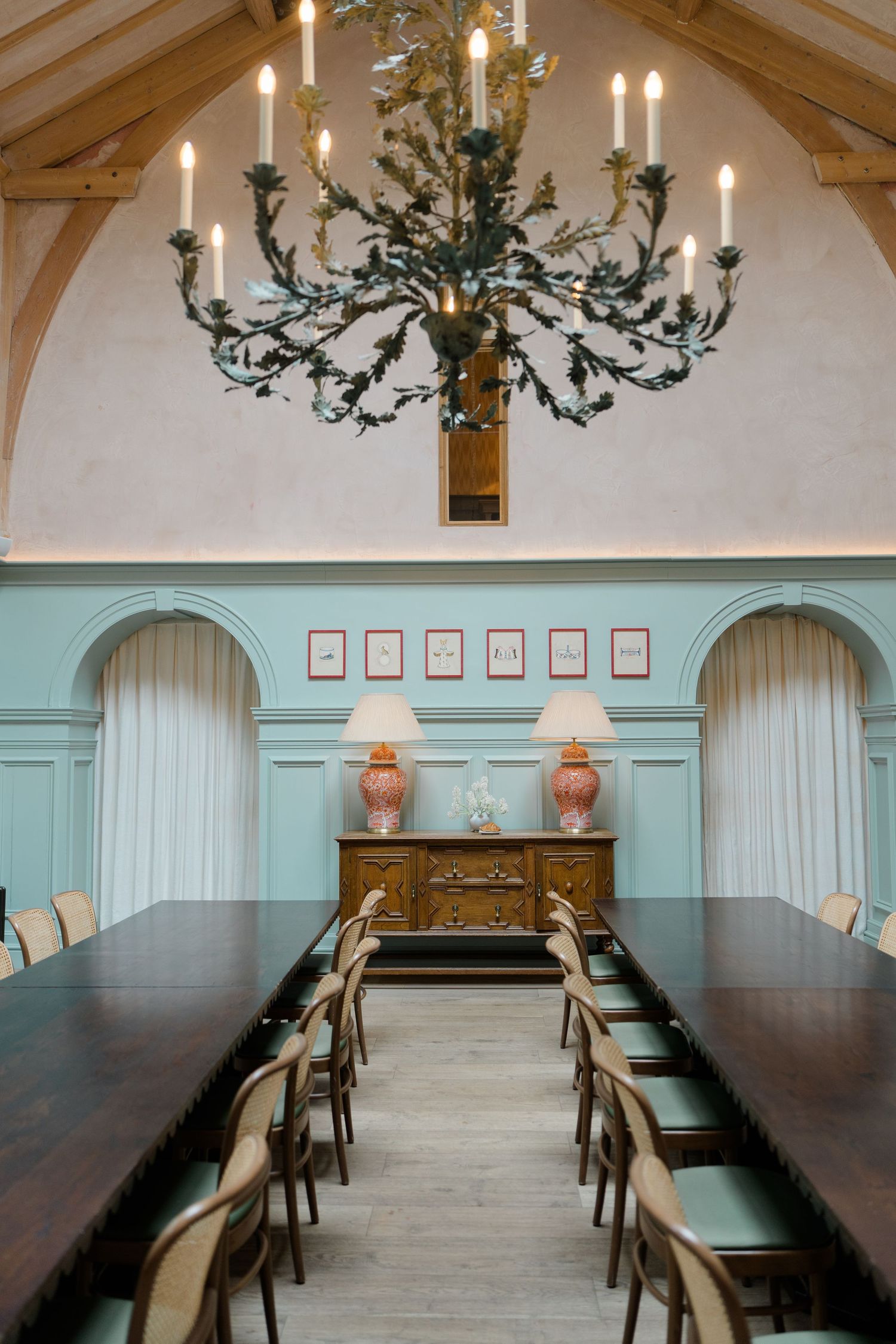 Elegant dining room with teal wainscoting, arched windows, long dark wood table, ornate chandelier, and antique sideboard with pink lamps.