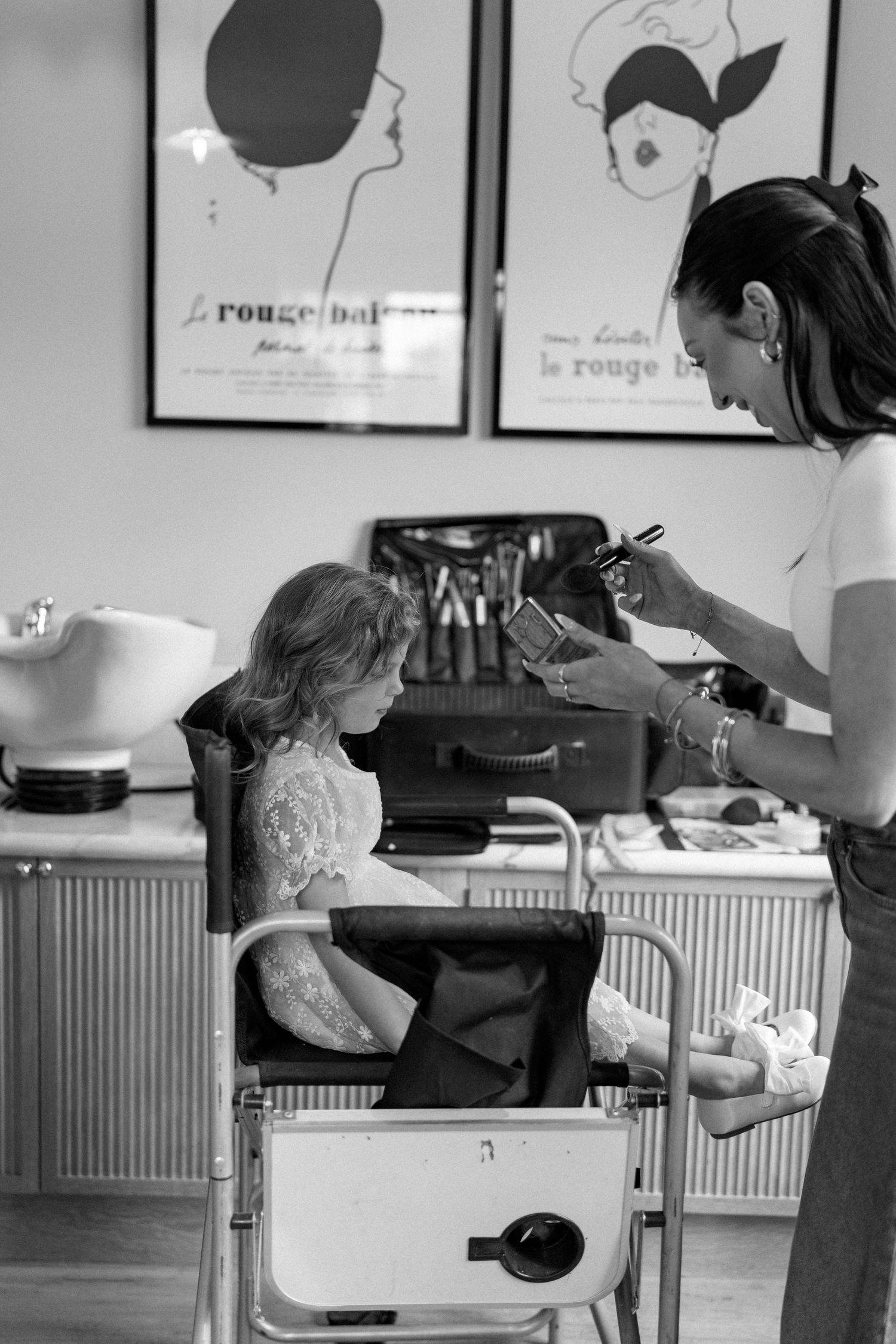 Black and white photo of young flower girl getting hair and makeup done in a bridal salon.