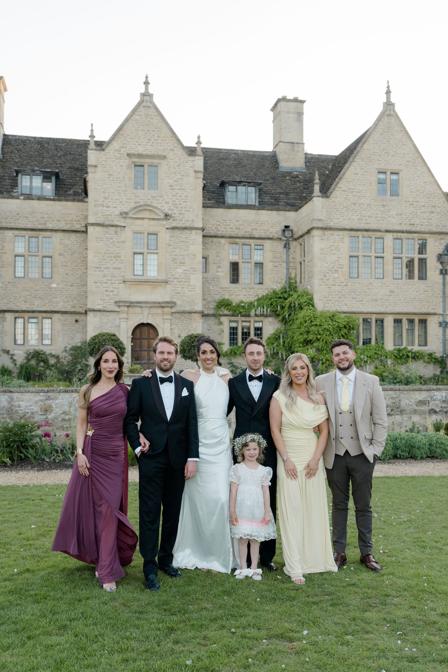 Wedding party poses before a grand stone manor house with bride, groom, and family members.