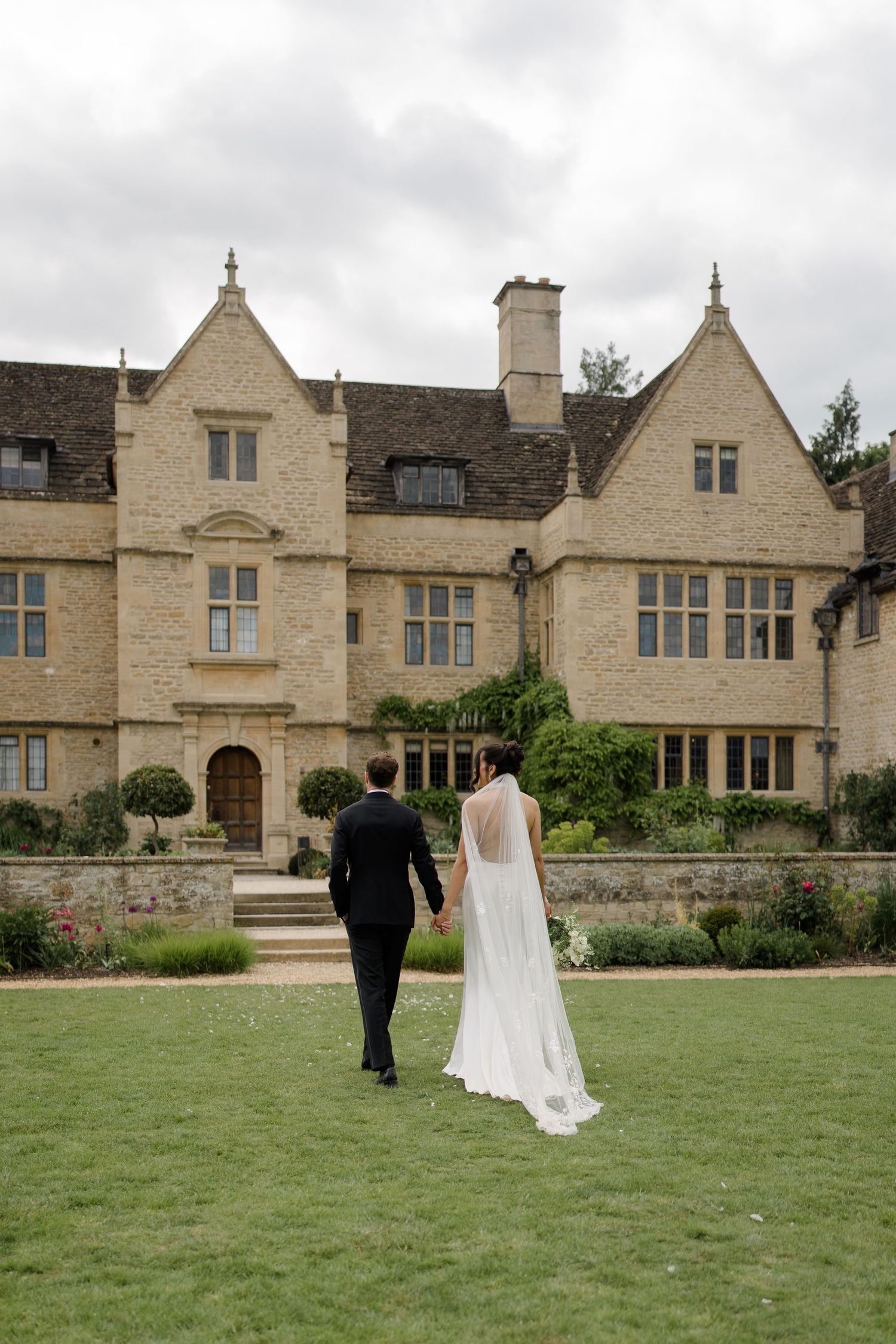Bride and groom walking hand in hand toward a grand English manor house on a cloudy day.