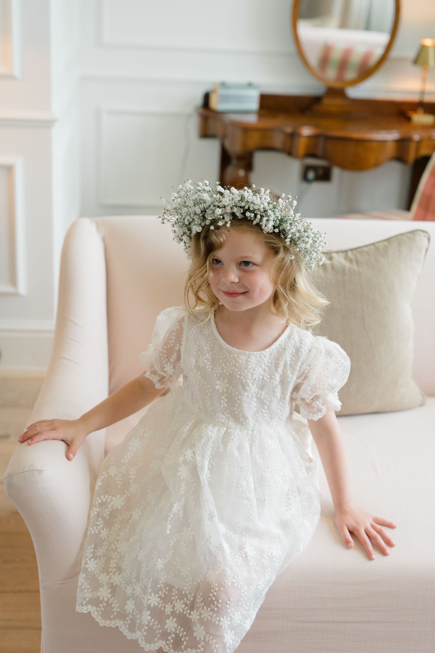 Adorable flower girl in white lace dress wearing a delicate baby's breath floral crown, posing by a cream armchair.