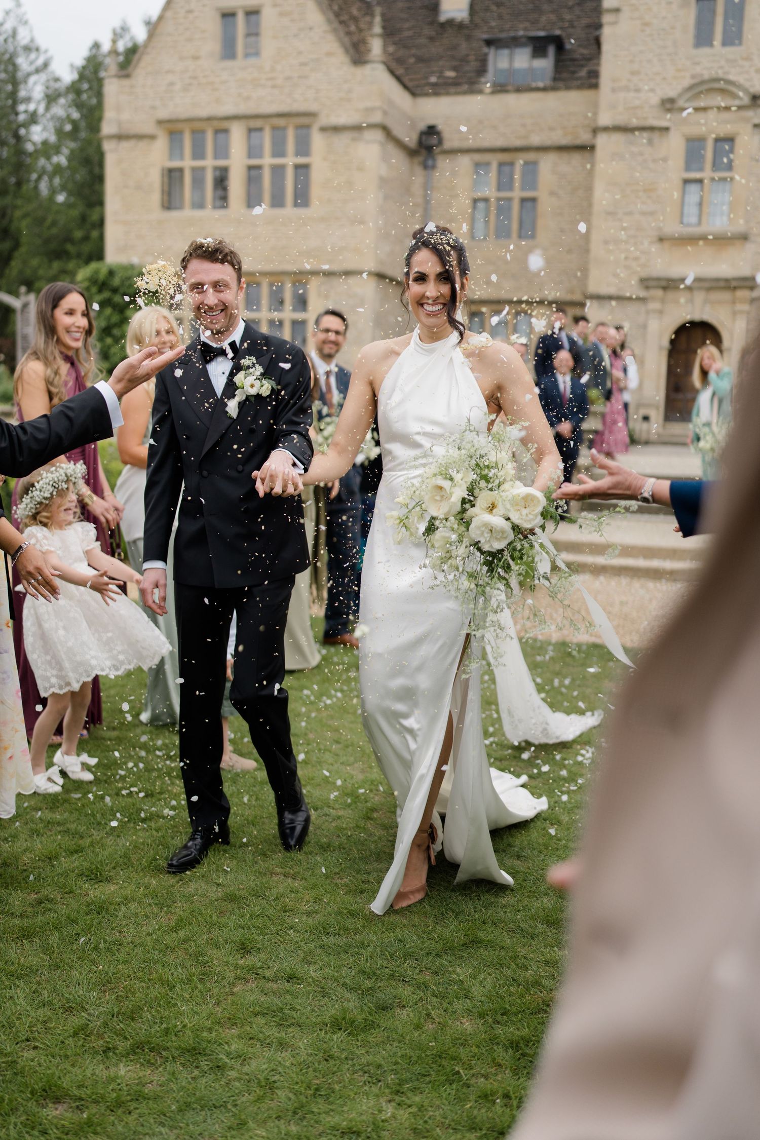 Bride and groom walk through confetti shower outside a stone manor house surrounded by smiling wedding guests.