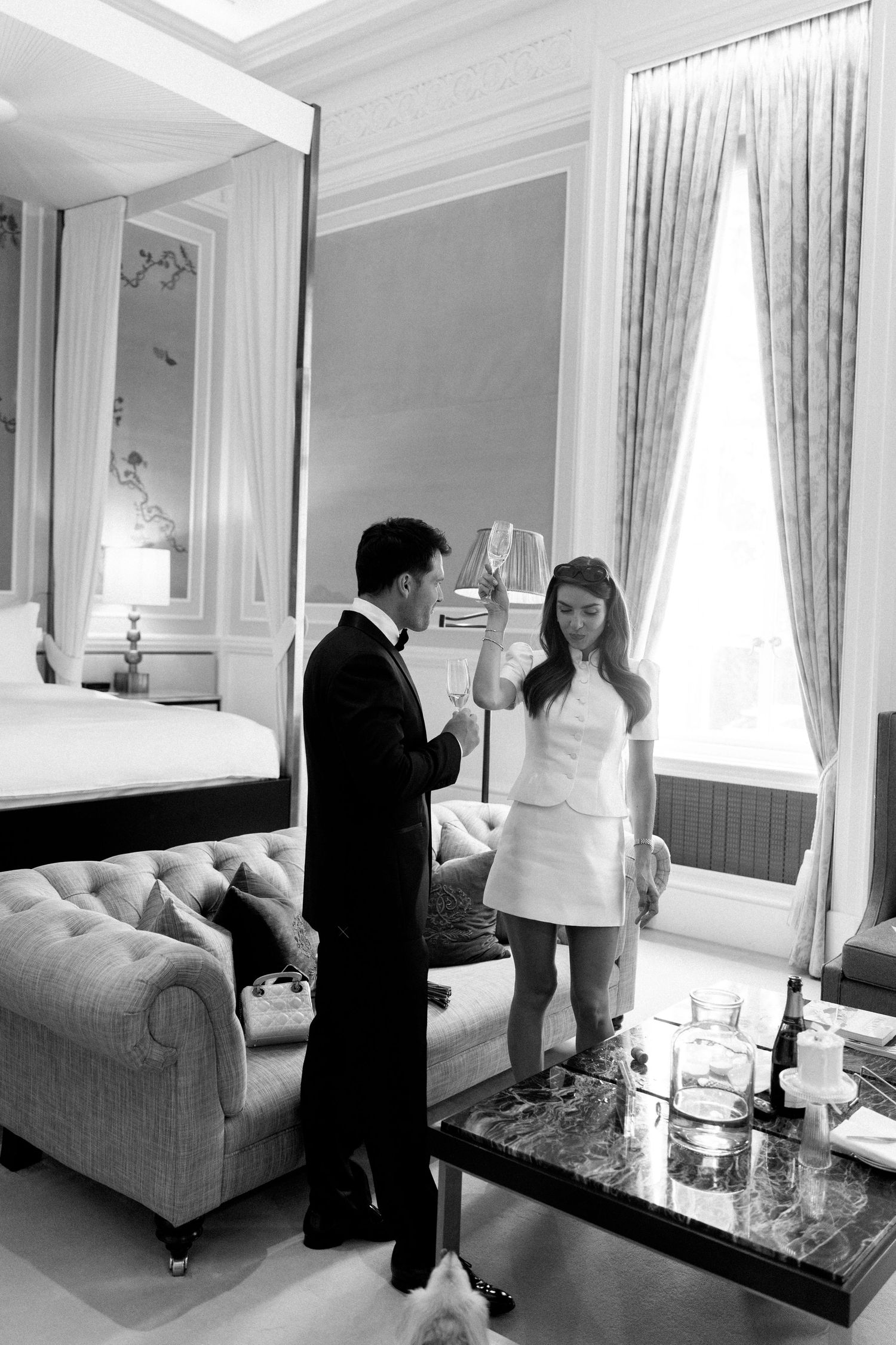 Black and white photo of bride and groom toasting with champagne in a luxurious hotel suite.